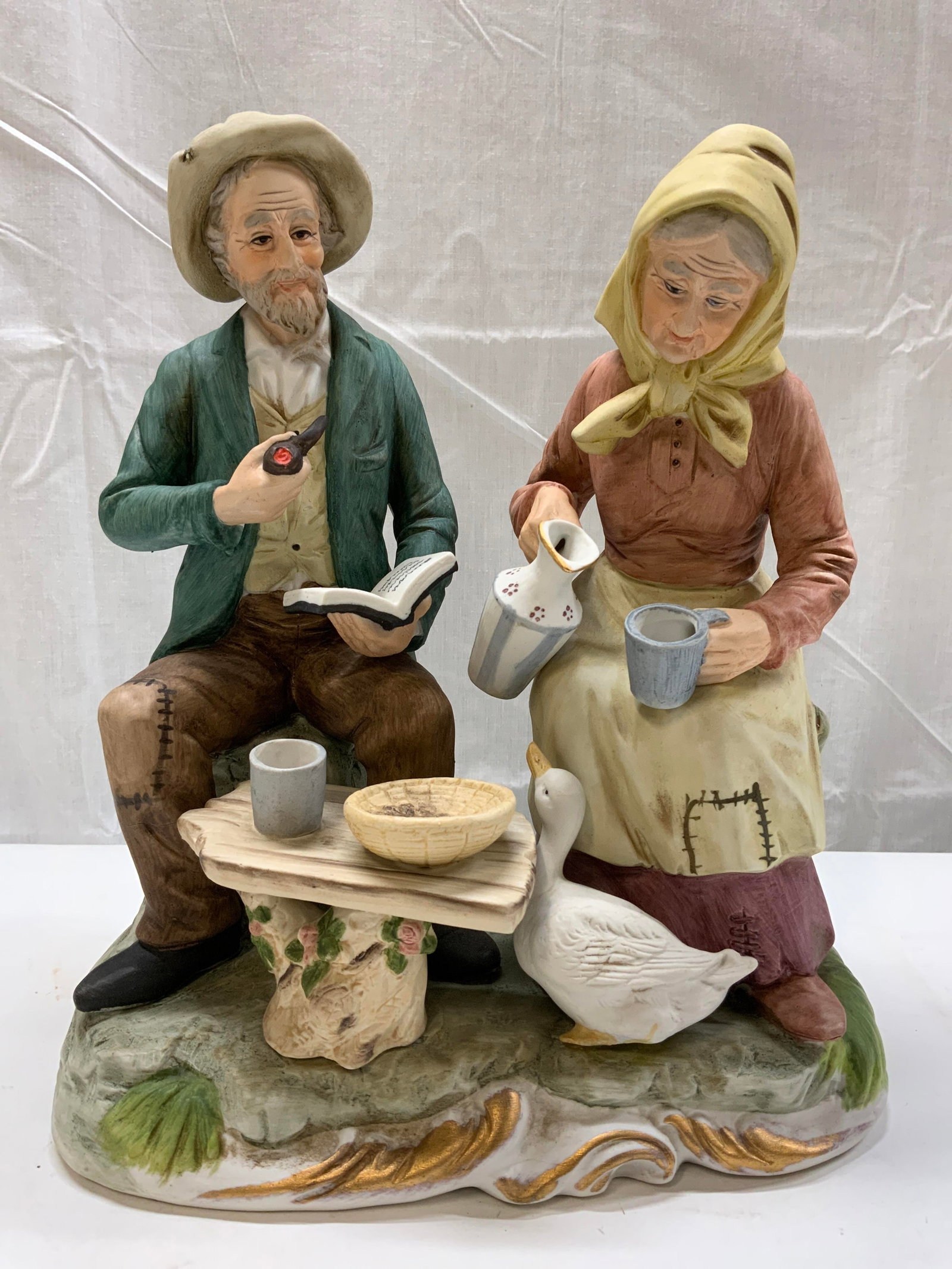 Vintage Porcelain Elderly Couple Figural: Vintage Porcelain Elderly Couple Figural Figurine of an elderly couple, elderly lady is holding a pitcher & mug. Elderly man is holding a pipe in one hand & and a book in the other. A white goose is s
