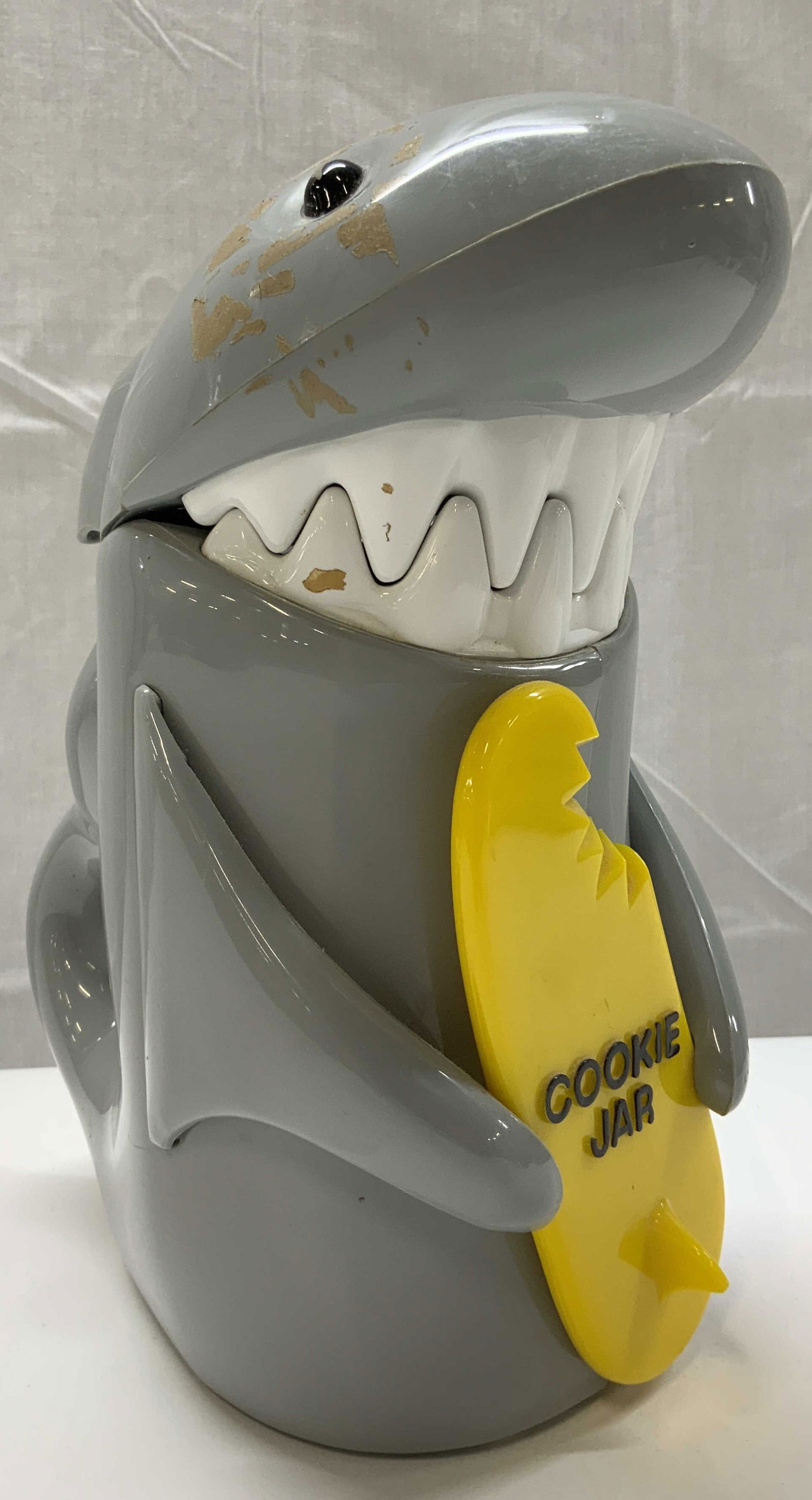 Vintage Grey Shark Cookie Jar: Vintage Grey Shark Cookie Jar Cookie jar in the shape of a grey shark. Shark is holding a yellow surf board that says COOKIE JAR. Shark has a tail in the back & big teeth. Cookie jar opens & has a slo