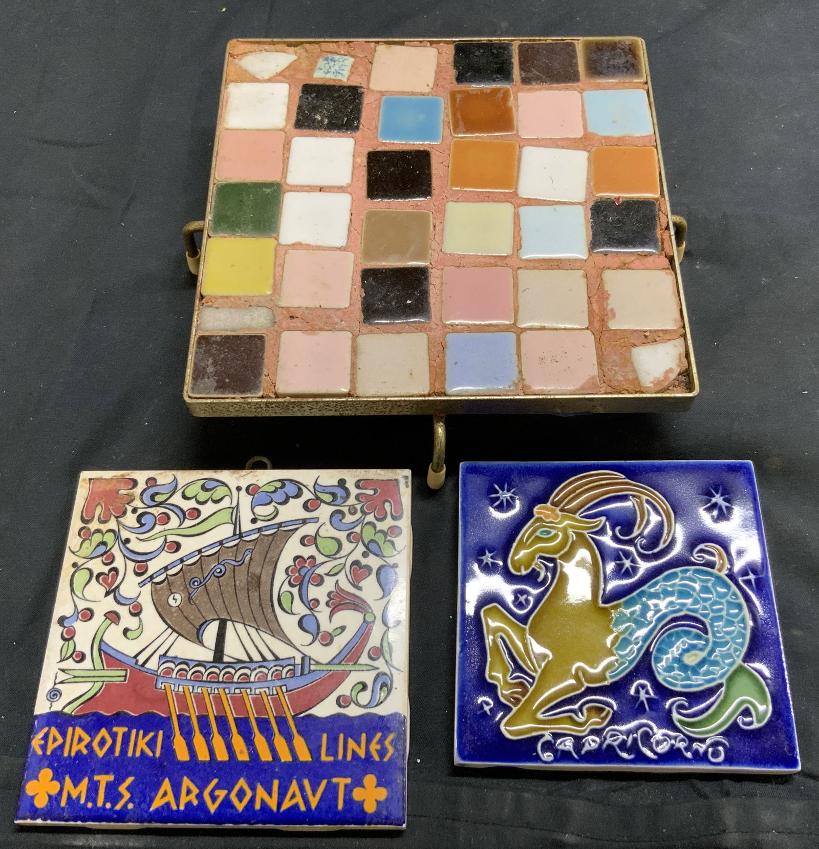 Lot 3 Vintage Porcelain Tile Trivets. Karos Crmcs+: Lot includes 1 mosaic trivet with footed metal base, 1 with ship floral and leaf detail (marked KAROS CERAMICS), & 1 Capricorn detail (Signed illegibly). Each piece appears to be vintage. Largest triv
