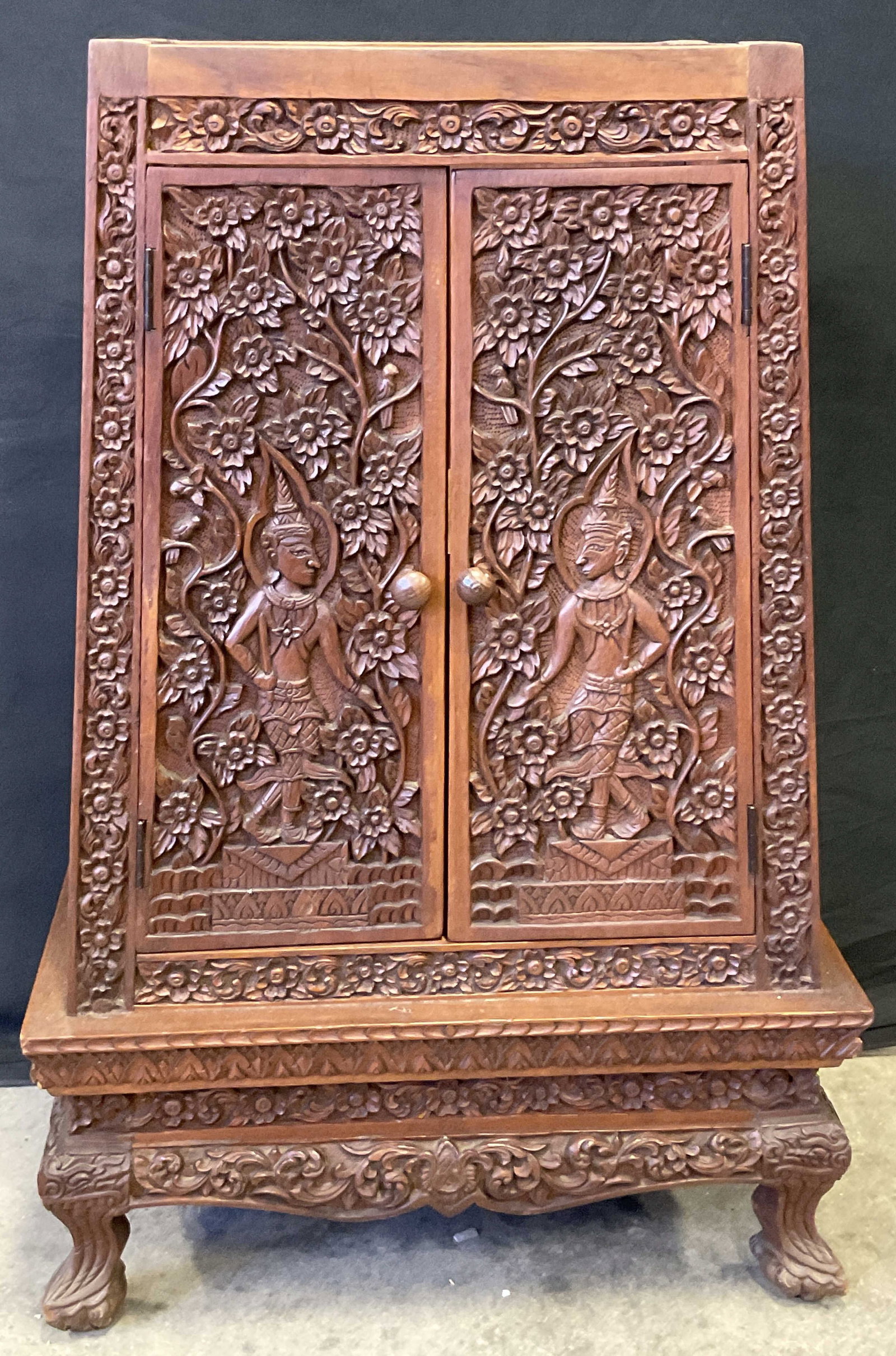 Vintage Asian Handcrafted Carved Cabinet: Vintage Asian handcrafted wooden cabinet featuring intricate carvings. The doors of the cabinet are adorned with detailed carvings of figures surrounded by floral and foliage patterns. The carvings co