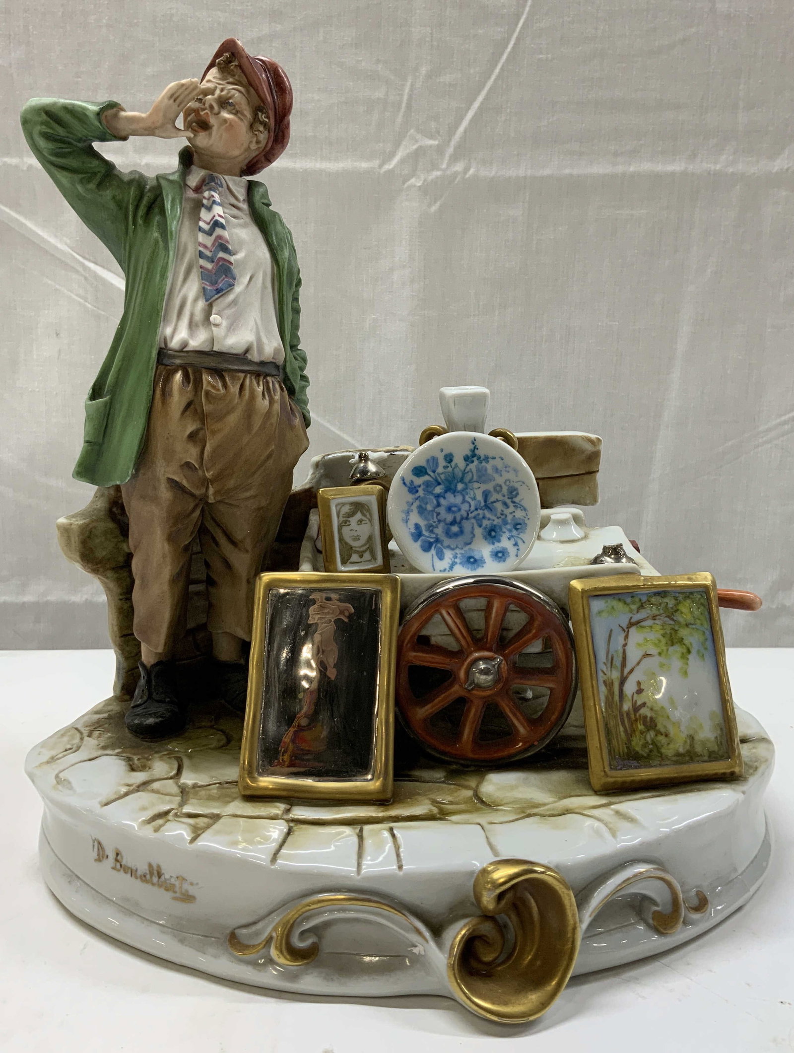 Vintage D.Bonathat Porcelain Figural w Cart ITALY: Vintage D.Bonathat Porcelain Figural w Cart ITALY Porcelain figural of a man in a green jacket, brown pants & a red cap. He is standing next to a cart with pictures frames, plates & more. The base has