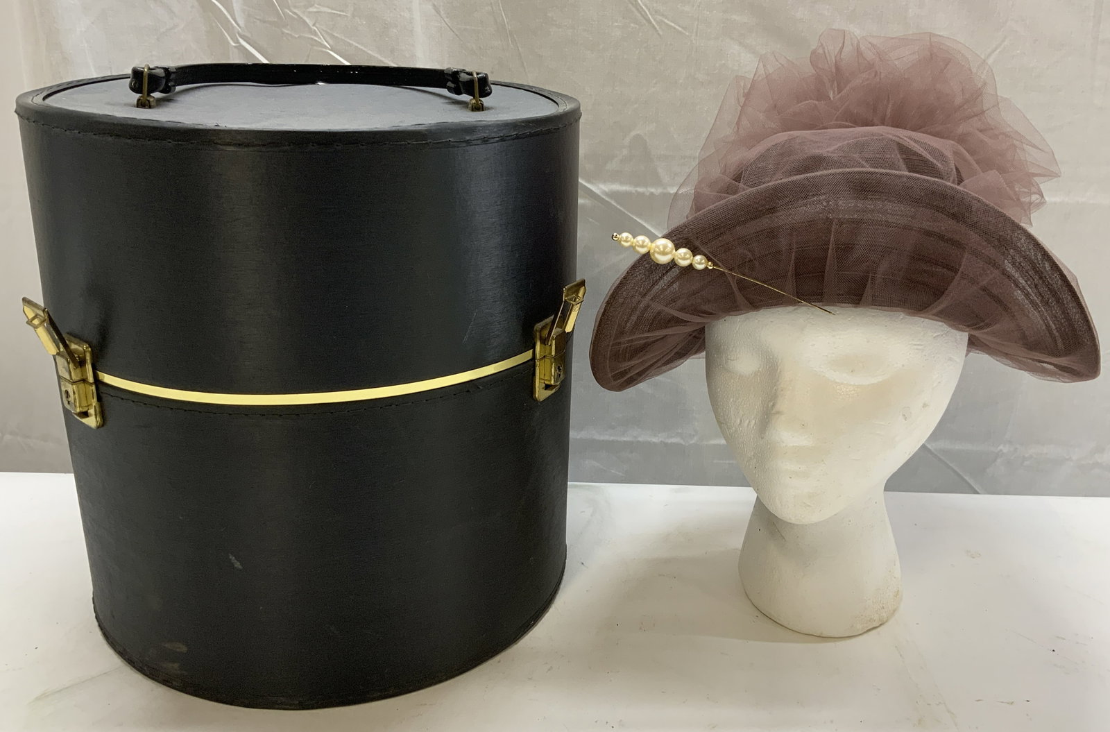 Vintage Purple Fascinator Hat w Black Round Case: Vintage Purple Fascinator Hat w Black Round Case Light purple fascinator hat that has a decorative pearl clip in the front. Hat comes with a black & gold round faux leather case. It has a handle & loc