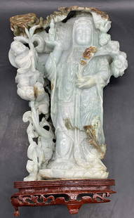 Carved Guan Yin Jade Sculpture W/ Wood Stand 14in: Carved Guan Yin Jade Sculpture With Wooden Stand. Figures are carved and etched amongst florals and foliage. The sculpture is attached to the stand. The stone is a light blue-green hue with dark green