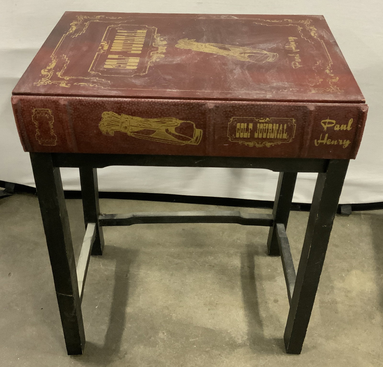 GOLF JOURNAL Paul Henry Wooden Side Table: GOLF JOURNAL Paul Henry Side TableSide table that has a book cover look as the tabletop. It has gold embossed lettering. GOLF JOURNAL PAUL HENRY. Tabletop has a maroon color & 4 black legs as the base