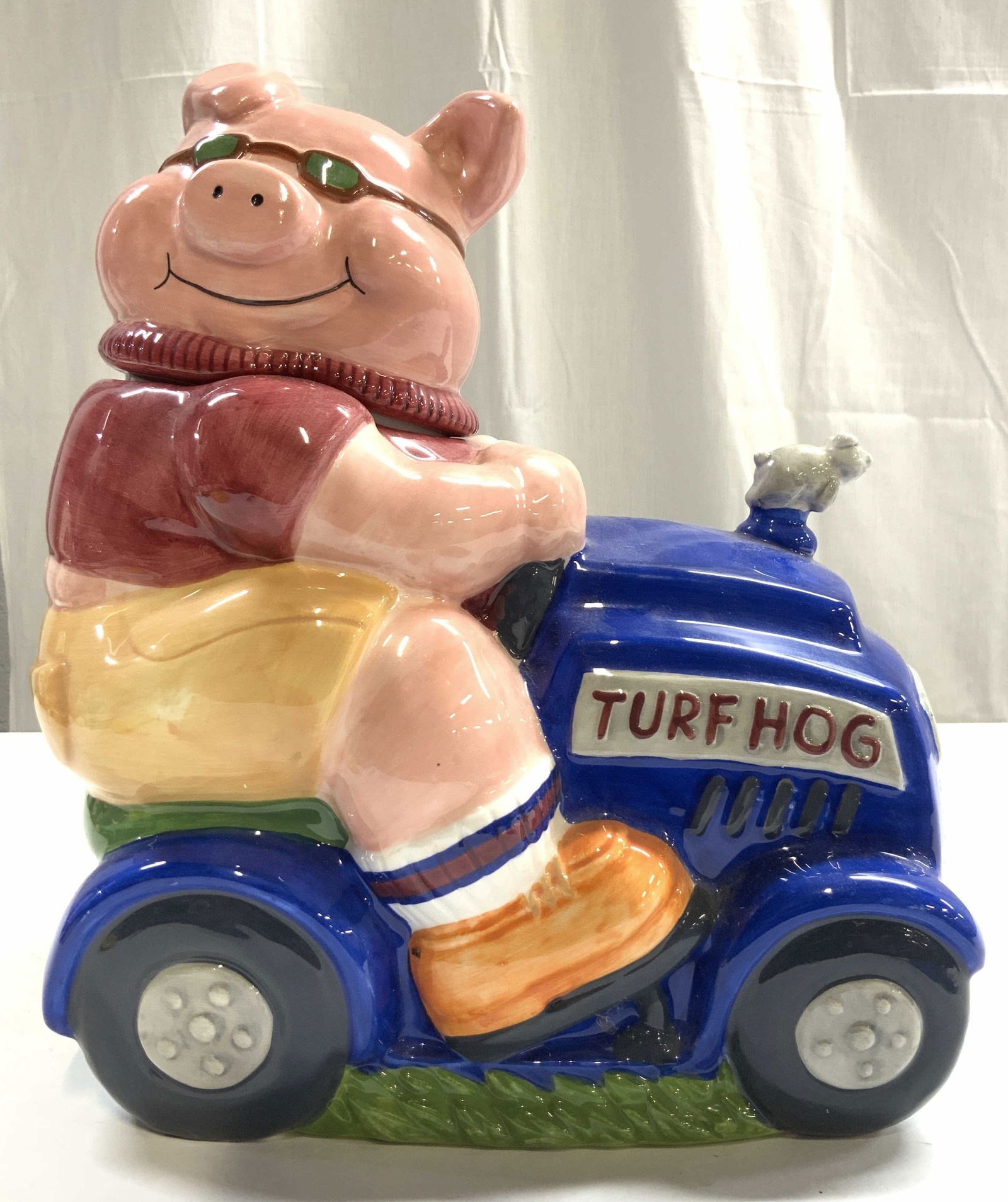 COOKS CLUB Porc Pig On Lawnmower Cookie Jar: Underside is marked COOKS CLUB. Cookie jar is porcelain. Piece is in the form of a pig on a lawn mover that reads TURF HOG. Turf has lid. Piece measures approximately 11.5 inches tall 10 inches deep 6