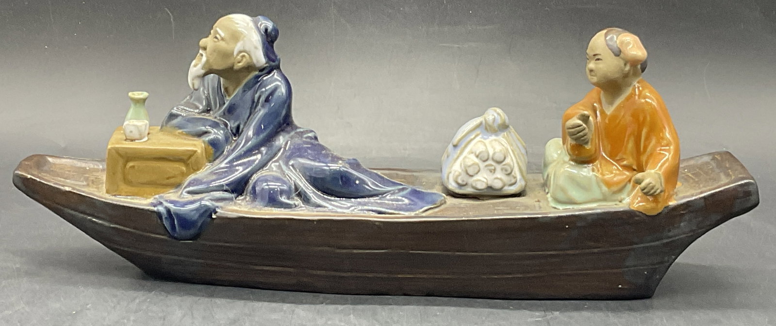 Vintage Asian Pottery Figures Atop a Boat Figural: Vintage Asian Pottery Figures Atop a Boat Figural. One person sitting towards the front of the boat in blue glazed clothing. The other person wears an orange glazed top and green hued pants. Measures 