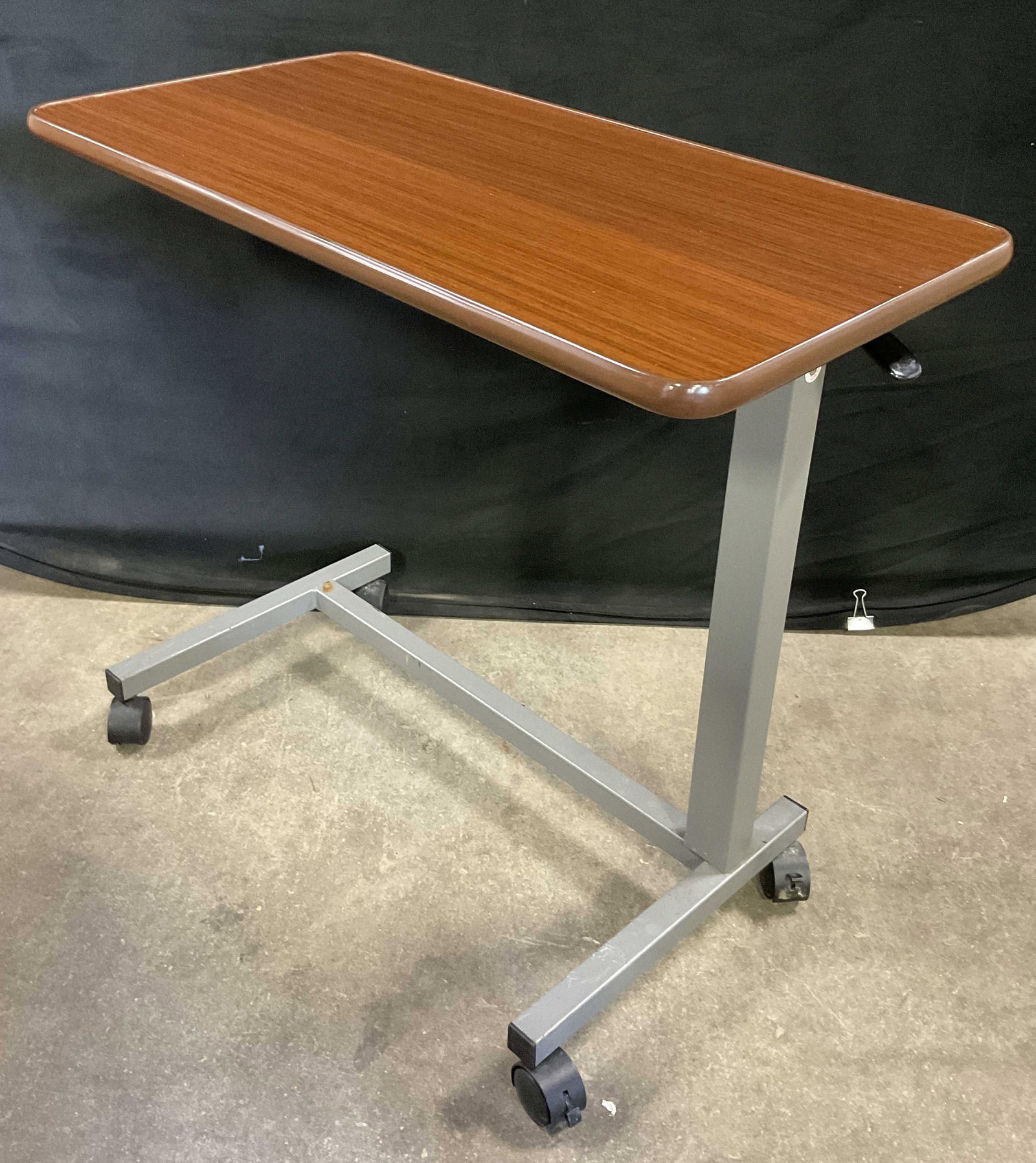 VAUNN MEDICAL Vintage Wheeled Medical Table: VAUNN MEDICAL Vintage Wheeled Medical Table. Rounded rectangular form. Brown hued pressed wood / veneer top and metal base. Wheels to the base. Measures approx. 30.25W x 15.5D x 28H in. Sticker reads 