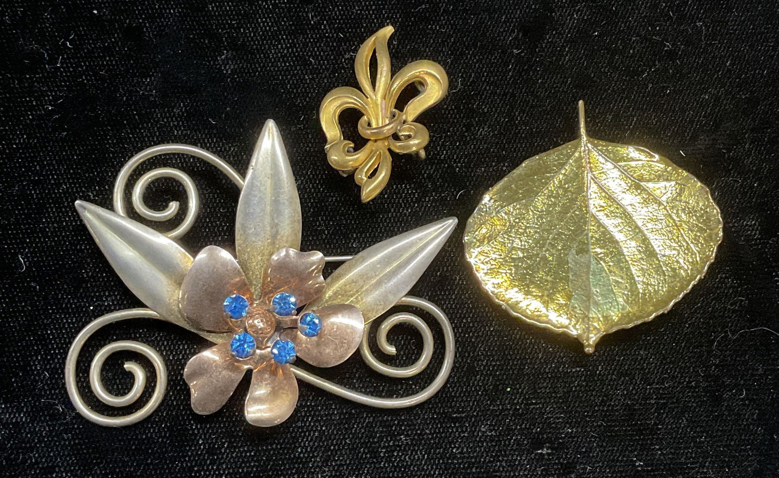 Lot 3 Vintage Gold Tone Botanical Brooches: Lot of 3 vintage gold tone botanical motif brooches. Blue Crystal, flower, leaf, fleur de lis. Fleur de lis brooch signed PSCO. Largest approx 1.75 x 2.25 inches. Some wear including scratching observ