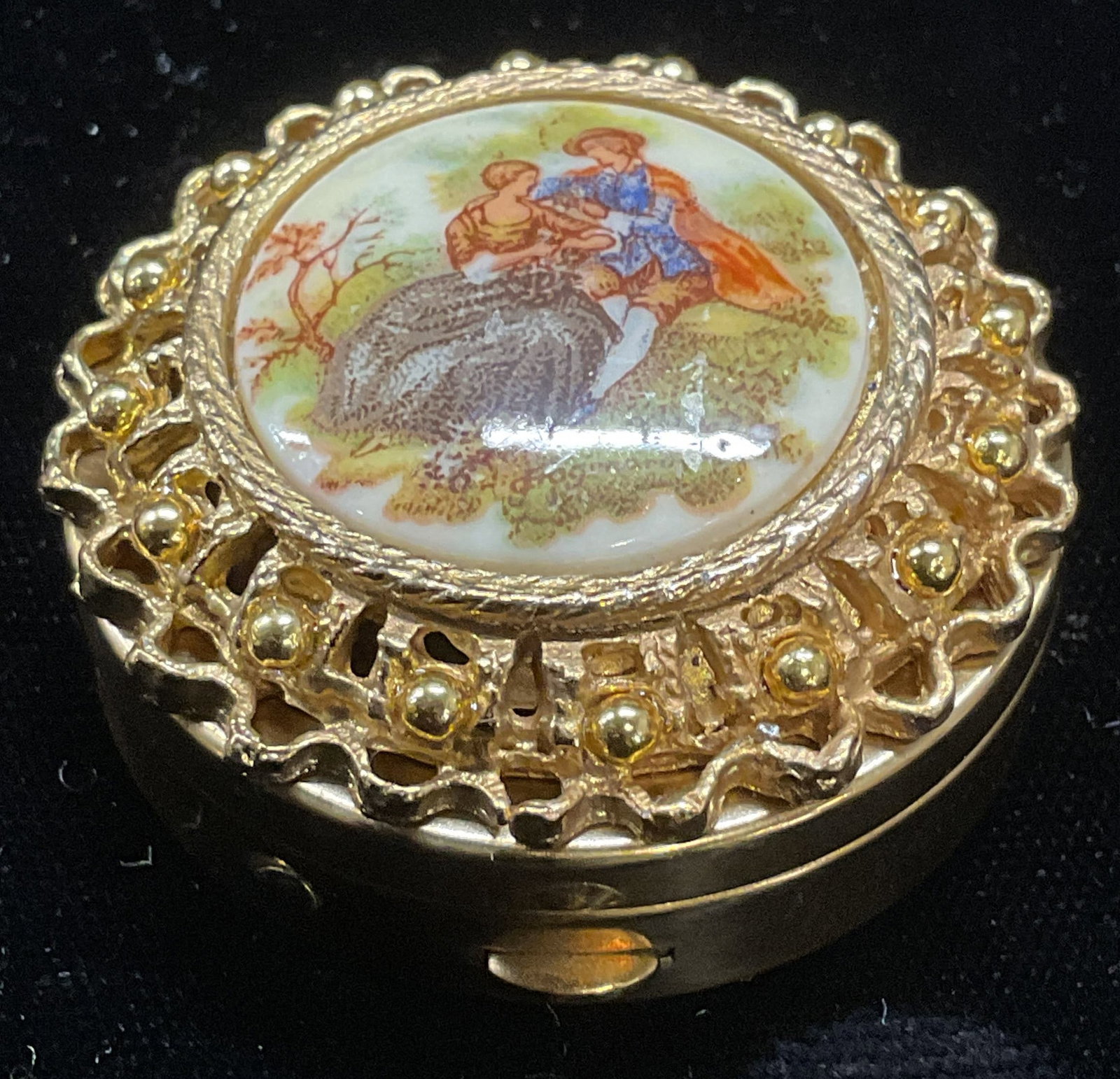 Vintage Gold Tone Couple Trinket Box: Vintage gold tone round trinket box with raised filigree design and courting couple motif. Hand painted decoration. Measures approx 1.5 x 1.25 inches. Some wear consistent with exposure. Inventory fro