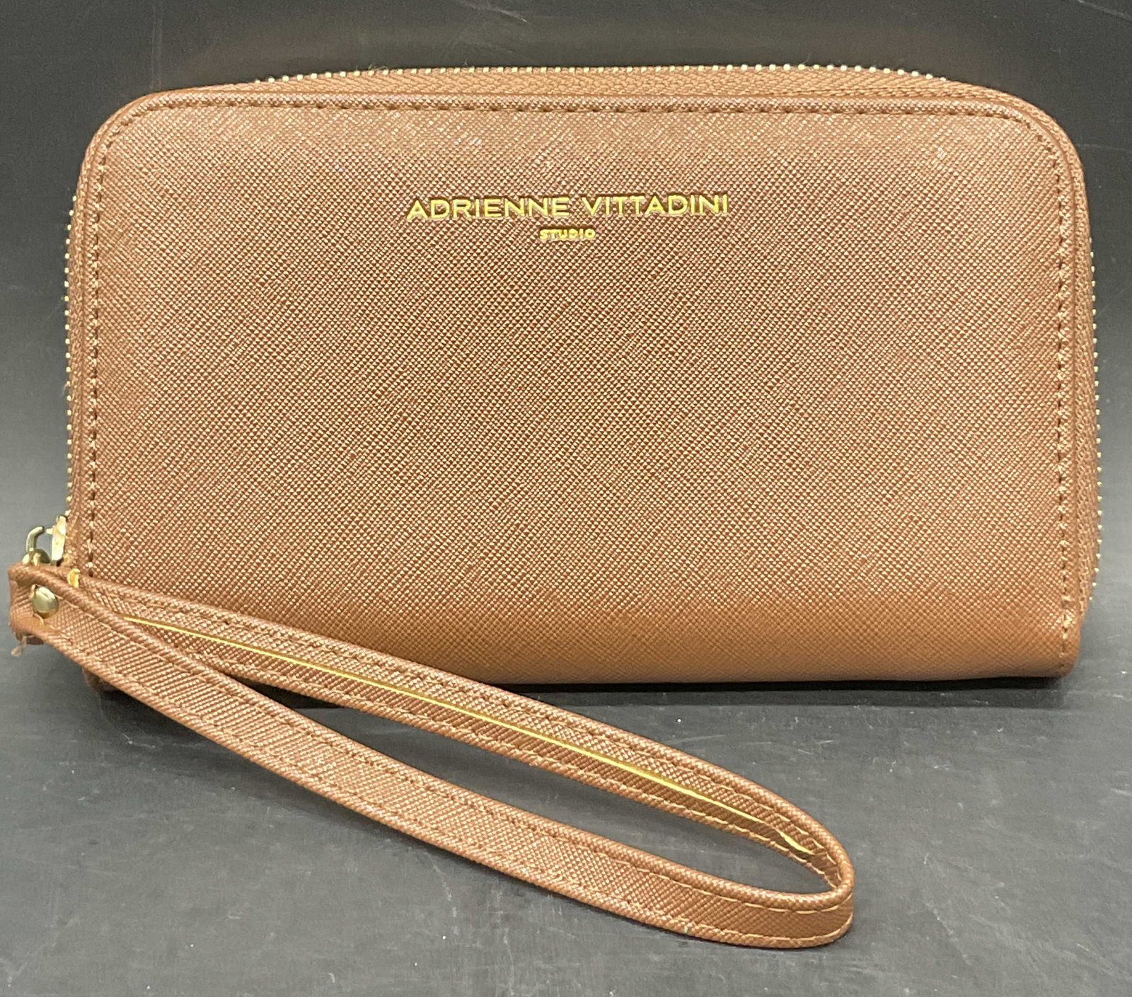 ADRIENNE VITTADINI STUDIO Brown Zippered Wristlet: ADRIENNE VITTADINI STUDIO Brown Zippered Wristlet. Obverse verbiage in gold tone letter reads ADRIENNE VITTADINI STUDIO. Measures approx. 7W x 4L in. Fabric lined interior with compartments for storag