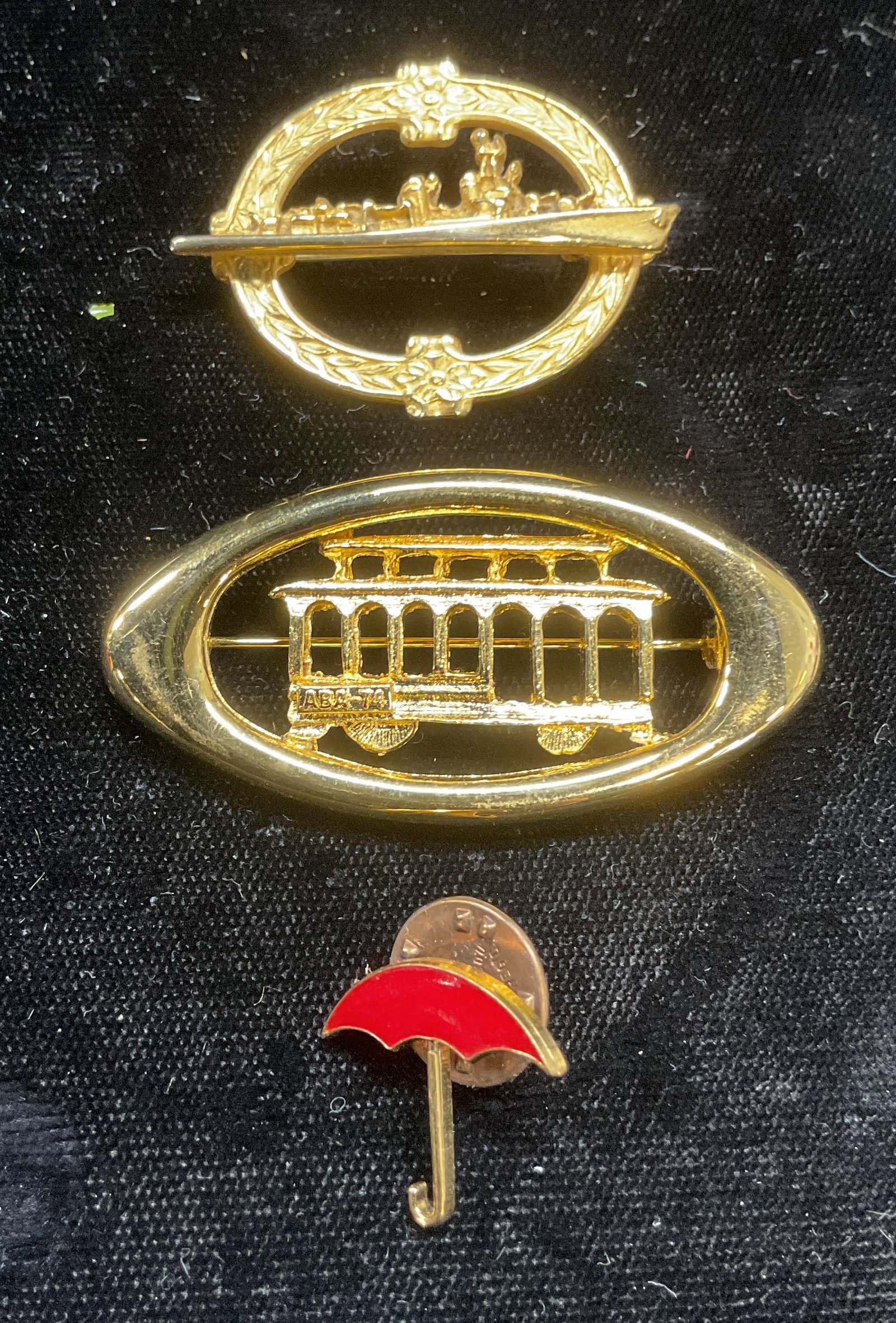 Lot 3 Trolley, Boat Brooches, Umbrella Pin: Lot of 3 gold tone brooches and pin. Trolley motif brooch, boat motif brooch, and red enamel umbrella motif pin. Largest measures approx 1 x 2 inches. Some wear including scratching observed. Jewelry,