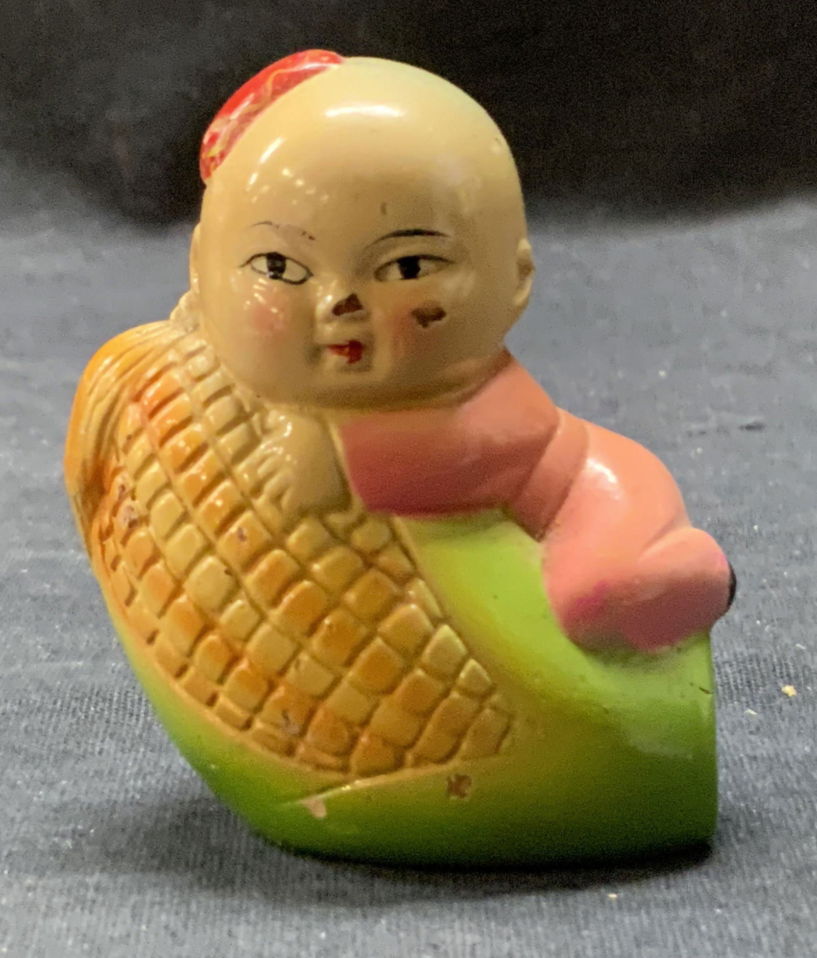 Vintage Child W/ Corn Figural Pencil Sharpener: A vintage pencil sharpener featuring a child sitting atop an ear of corn. Piece is crafted from composite, ceramic, and metal materials. Measures approx. 2.5H x 2W in. Chipping, losses to finish, and