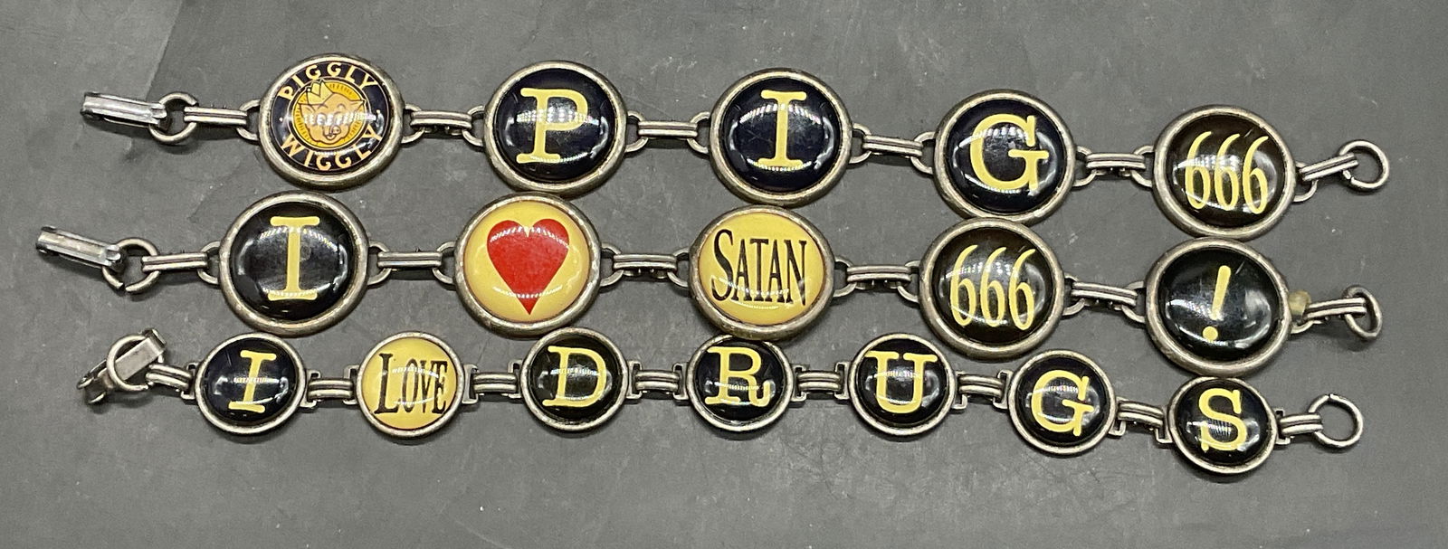 Lot 3 Phrase Charm Bracelets, PigglyWiggly+: Three Phrase Charm Bracelets. Pieces secure with metal clasp closures. One reads I Love Drugs. Another reads Pig, 666, and Piggly Wiggly. Pieces are similar in length measuring approx. 7.75 inches end