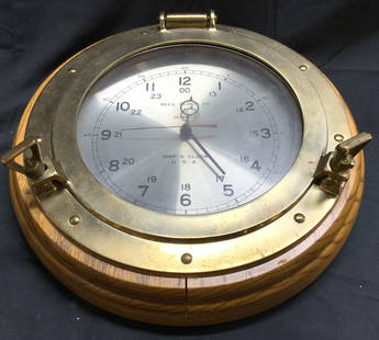 BELL CLOCK CO Wood Veneer & Brss Porthole Clk: Clock face reads BELL CLOCK CO. Clock frame is tan toned wood veneer and brass. Piece has brushed metal silver toned face with black toned lettering and numbers. Porthole clock measures approximately