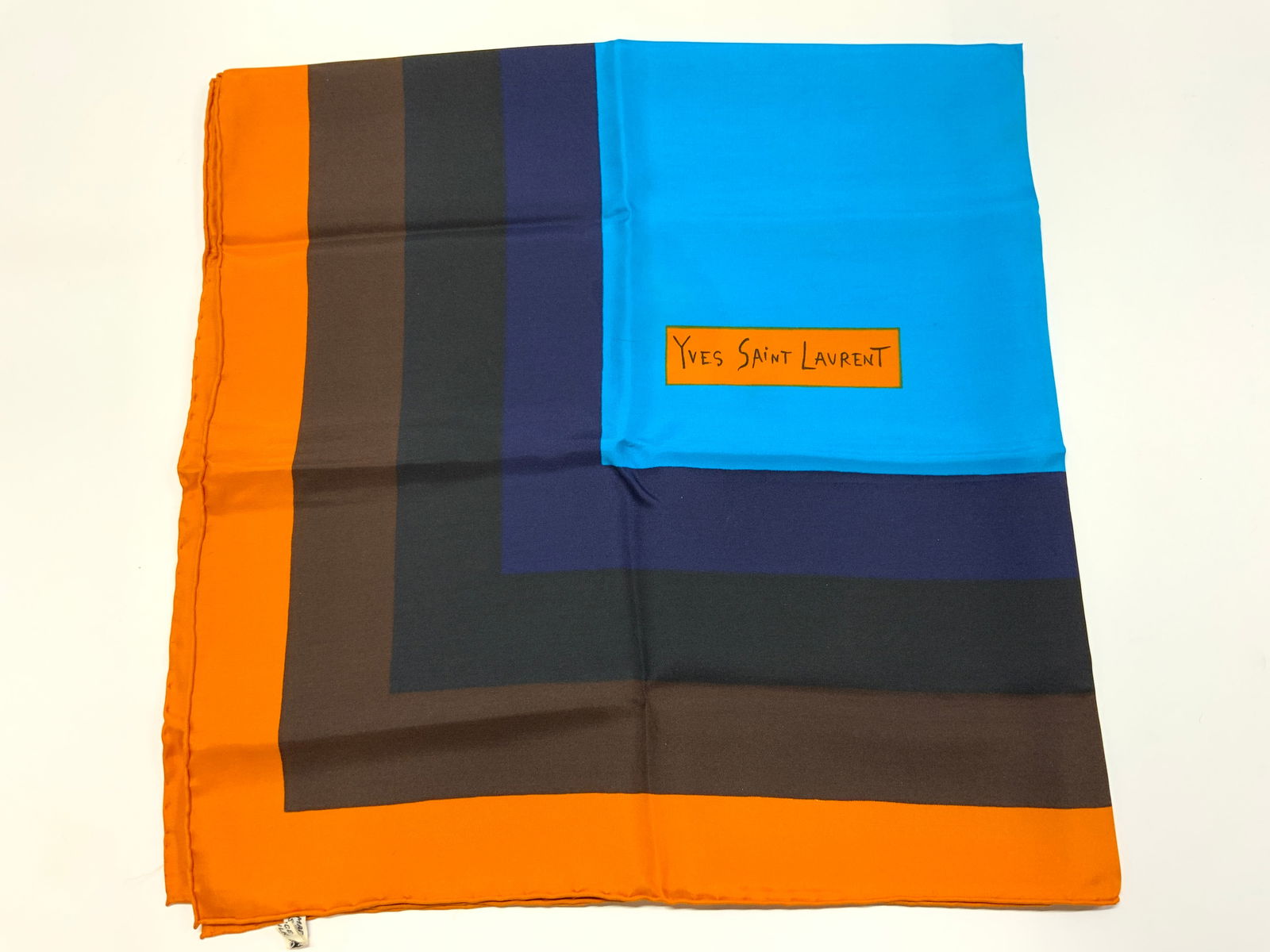 Yves Saint Laurent Silk Geometric Block Scarf France: Yves Saint Laurent Silk Geometric Block Scarf Silk scarf featuring a geometric & color-block design. Scarf has shades of blue, black, brown & orange. Silk scarf has YVES SAINT LAURENT text. MADE IN FR