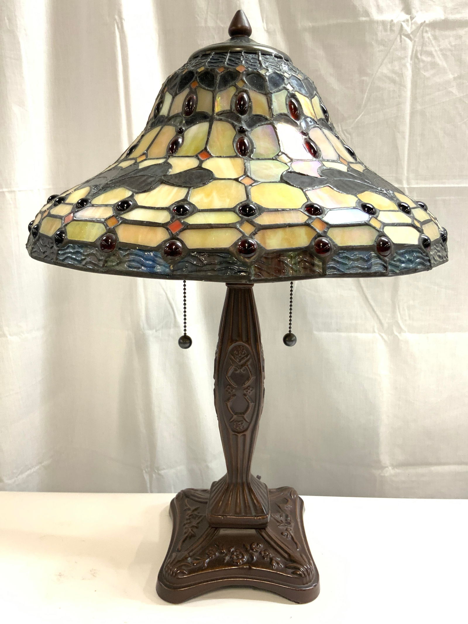 Tiffany Style Stained Glass Shade Table Lamp: Lamp is Tiffany Style. Piece has multi toned stained glass shade. Lamp has bronze toned metal base with floral and leaf detail. Piece has 2 beaded metal pull switches. Lamp includes metal finial. Piec