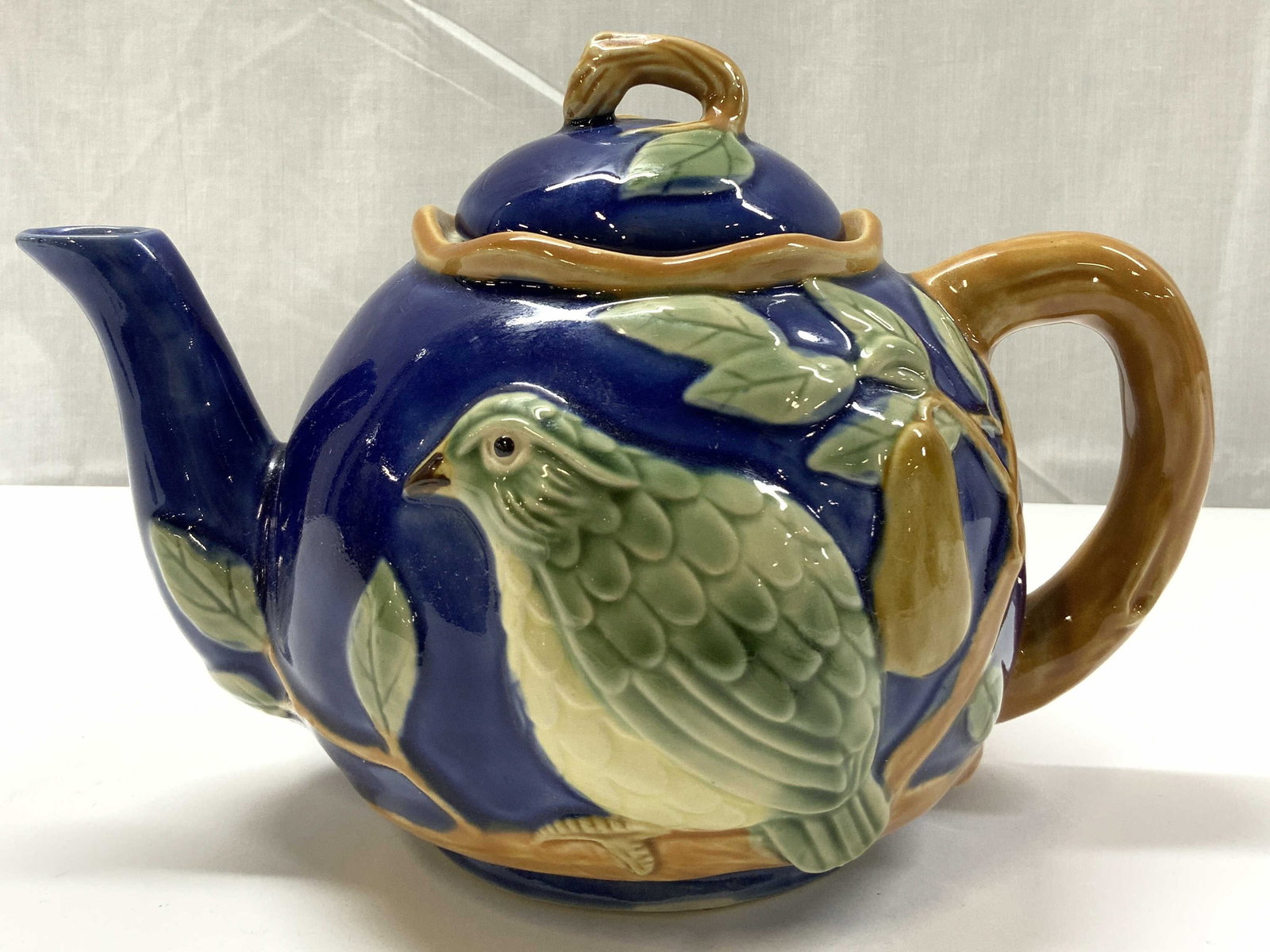Majolica Blue Partridge in a Pear Tree Teapot: Teapot depicting a green partridge in a pear tree. Teapot has a cobalt blue color & the branch of the tree forms as the handle. Surface wear consistent to age, use & exposure. Measurements: length 9.5