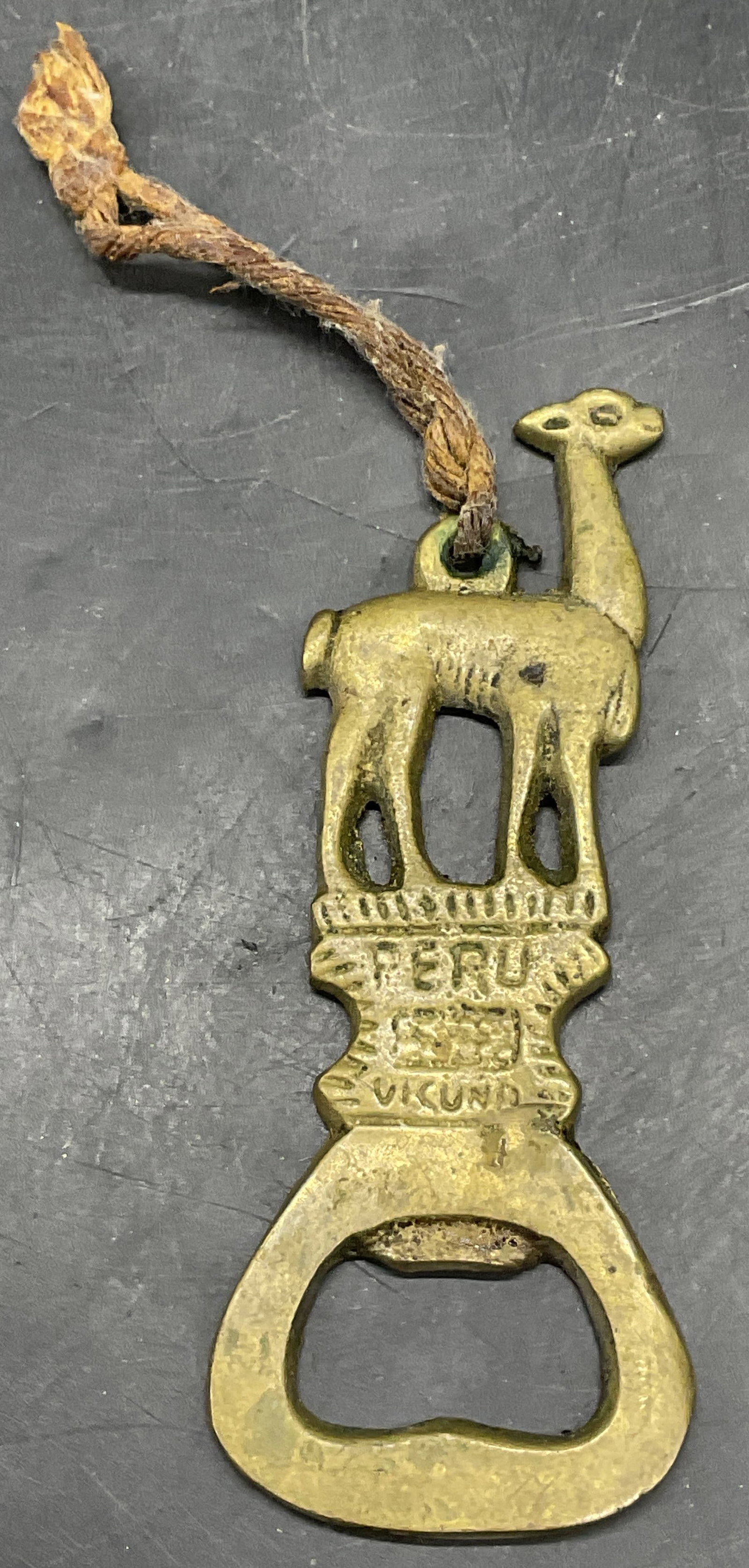 Brass Llama Motif Bottle Opener, Peru: Brass Llama Motif Bottle Opener. Obverse reads Peru. Other verbiage illegible. Measures approx. 3.5L x 1.5W in. Cord tied to the top. Discoloration, surface wear, and residue present. home decor, home