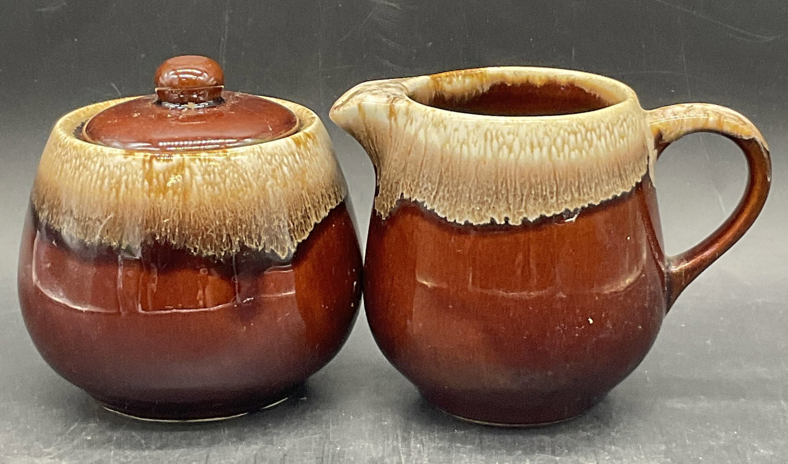 Pair MCCOY USA Pottery Creamer & Sugar Bowl: Pair of MCCOY USA Pottery Creamer & Lidded Sugar Bowl. Underside signed MCCOY USA 7020. Creamer measures approx. 4H x 5W in. Primarily brown and creamer hues. Drip glaze to the pieces. Discoloration, 