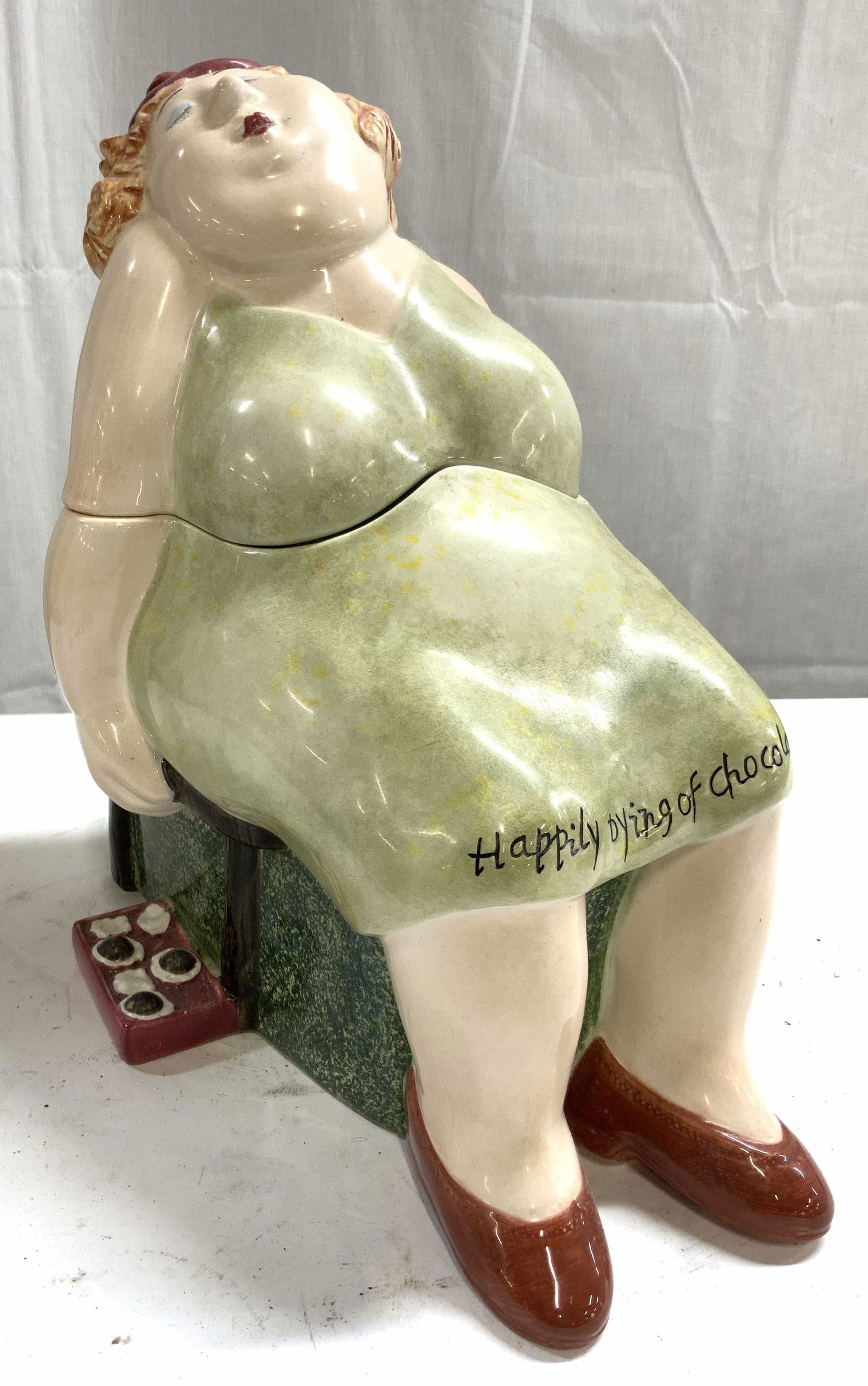 ERIKA OLLER HOUSE OF PRILL Fig Prc Cook Jar 2002: Underside is marked ERIKA OLLER HOUSE OF PRILL. Cookie jar is porcelain. Piece depicts a seated female figure wearing a dress which reads HAPPILY DYING OF CHOCOLATE. Cookie jar includes lid. Piece mea