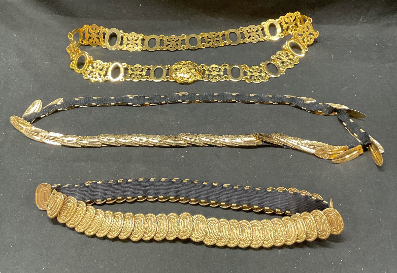 Lot 3 Vtg Gold Tone Metal and Black Elastic Belts: Lot of 3 Vintage Gold Tone Metal & Black elastic belts. Longest Belt length approx. 36 inches. Hook closure. Black material appears to be elastic. Losses, surface wear, and discoloration present. Wear