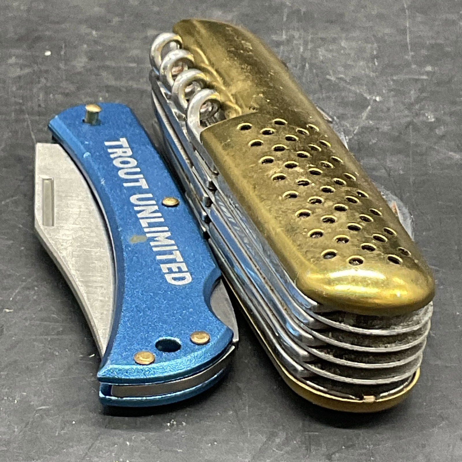 Lot 2 Metal Pocket Knives, TROUT UNLIMITED +: Two metal pocket knives. One piece features blue hued accents and is marked TROUT UNLIMITED, and the other features gold tone metal accents. Larger pocket knife measures approx. .75H x 3.25W in. Disco