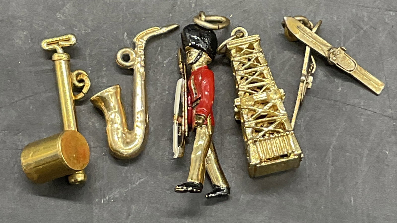 Lot 5 Vintage Gold Tone Metal Charms, Soldier +: Five Vintage Gold Tone Metal Charms. Charms include a soldier, skis, a building, and more. The underside of the building is marked AG or AC, worn. The building measures approx. .25W x 1L in. Discolora