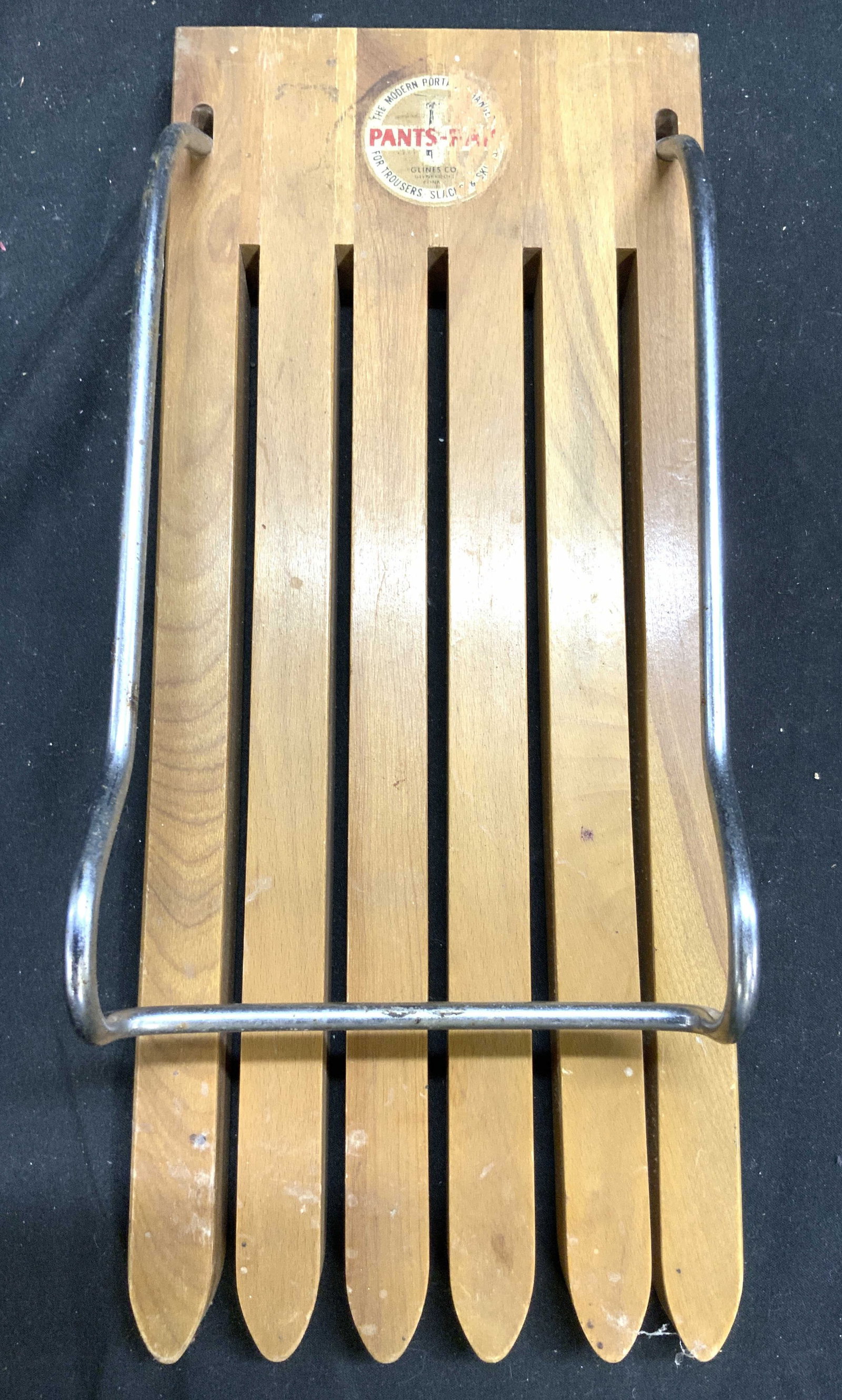 GLINES CO Vntg Wood & Metal Pants Rack: Tag reads GLINES CO. PANTS RAK. Pants rack is tan toned, appears to be wooden (possibly wood veneer) and metal. Vintage Wall Mount pants Rack with toothed lower area. Metal structure is adjustable. Pi