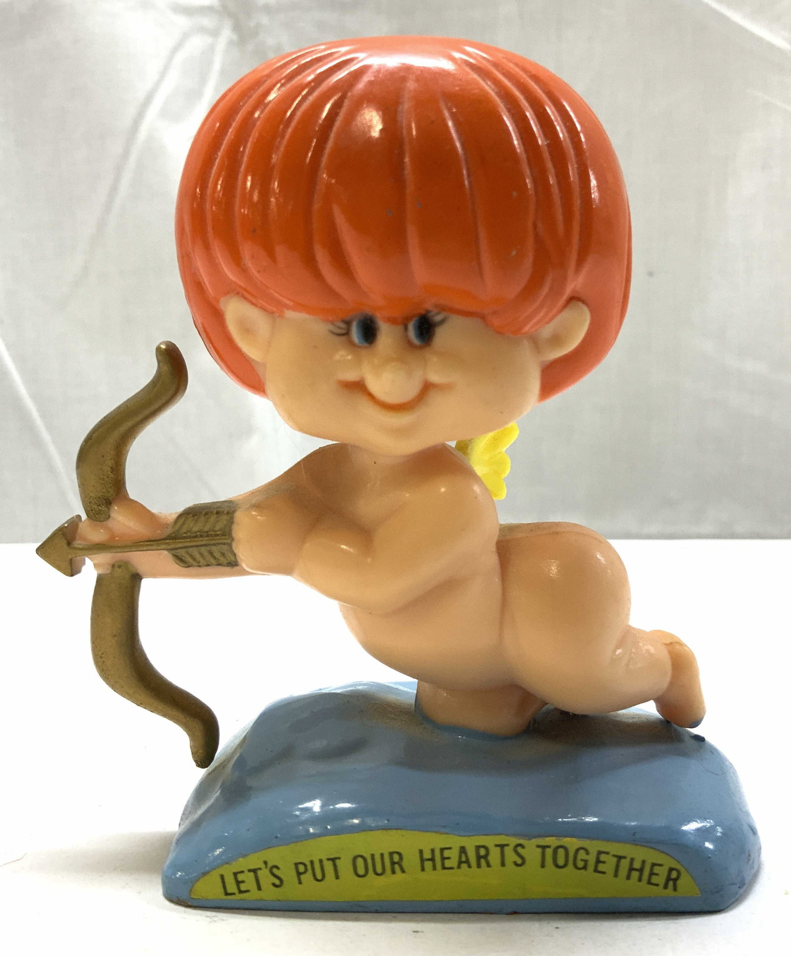 1971 RUSS BERRIES Red Cupid Figurine HONG KONG: 1971 RUSS BERRIES Red Cupid Figurine HONG KONG Figurine depicting a Cupid with red hair holding a bow & arrow, positioned on a base with text that reads LETS PUT OUR HEARTS TOGETHER. Surface wear cons