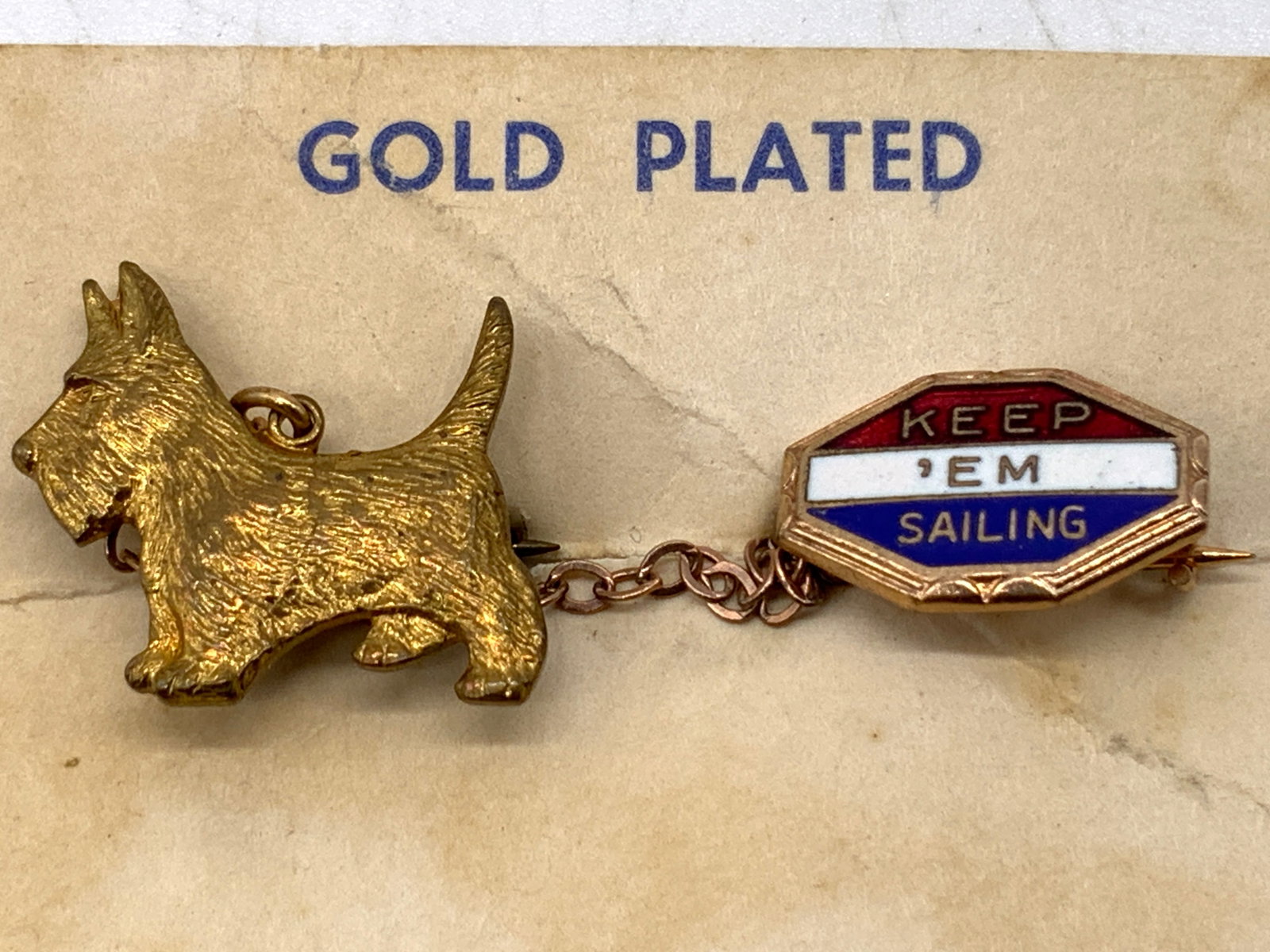 Gold-Plated Sailboat & Scottie Dog Chain Pin: Gold-Plated Sailboat & Scottie Dog Chain Pin Pin features a Scottie dog attached to a chain leading to a dangling emblem. Emblem reads KEEP EM SAILING. Surface wear consistent to age, use & exposure. 