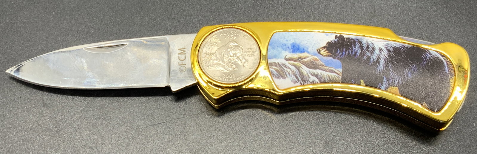 Stainless Steel Folding Knife W Alaska Bear Coin: Stainless Steel Folding Knife W Alaska Bear Coin Folding knife features an image of a black bear & an embedded coin. Coin has an illustration of a bear eating a fish. Knife has a bright gold-toned col