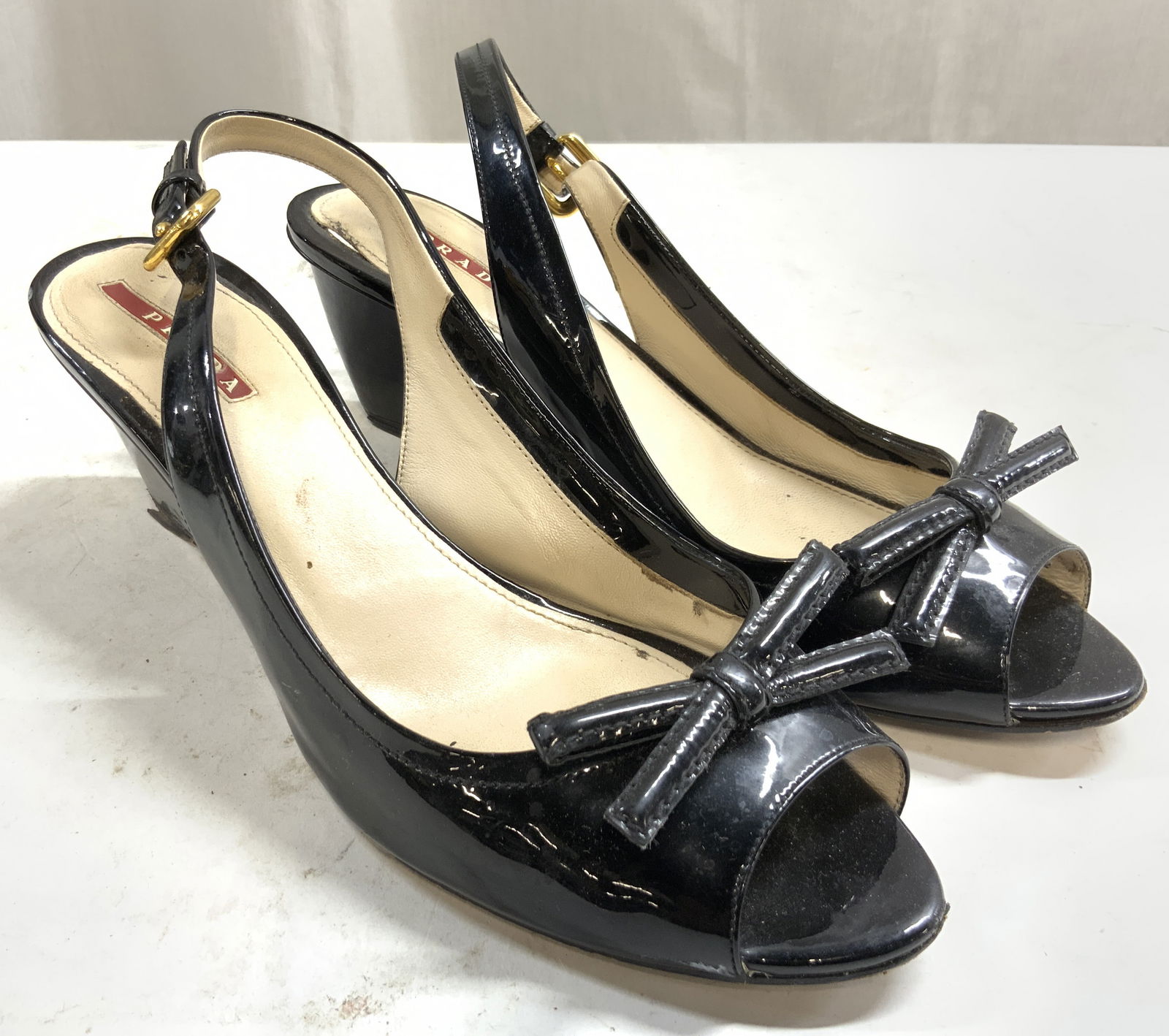 PRADA Patent Leather Heels Blk 39.5 ITALY: Heel sole reads PRADA. Underside is inscribed MADE IN ITALY. Heels are black toned patent leather sole is cream toned leather underside size reads 39 1/2. Heels have bow form shoe ornament. Peep toe h