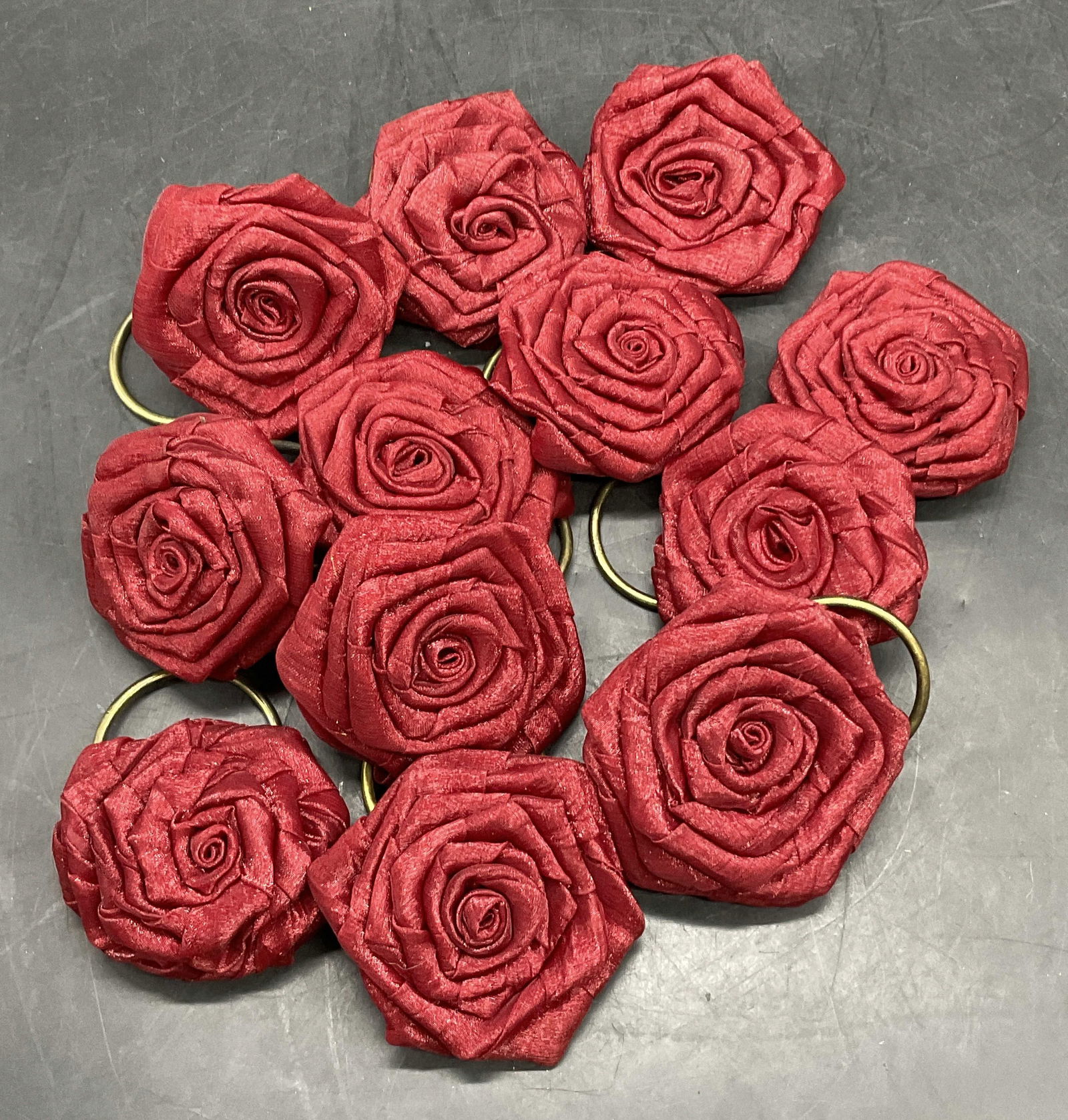 Set 12 Red Floral Rosette Napkin Rings: Set of 12 Red Floral Rosette Napkin Rings. Crafted from fabric and metal. Rosettes are similar in size measuring approx. 2.5L x 2.5W in. Warping, surface wear, loose pieces, and discoloration present.