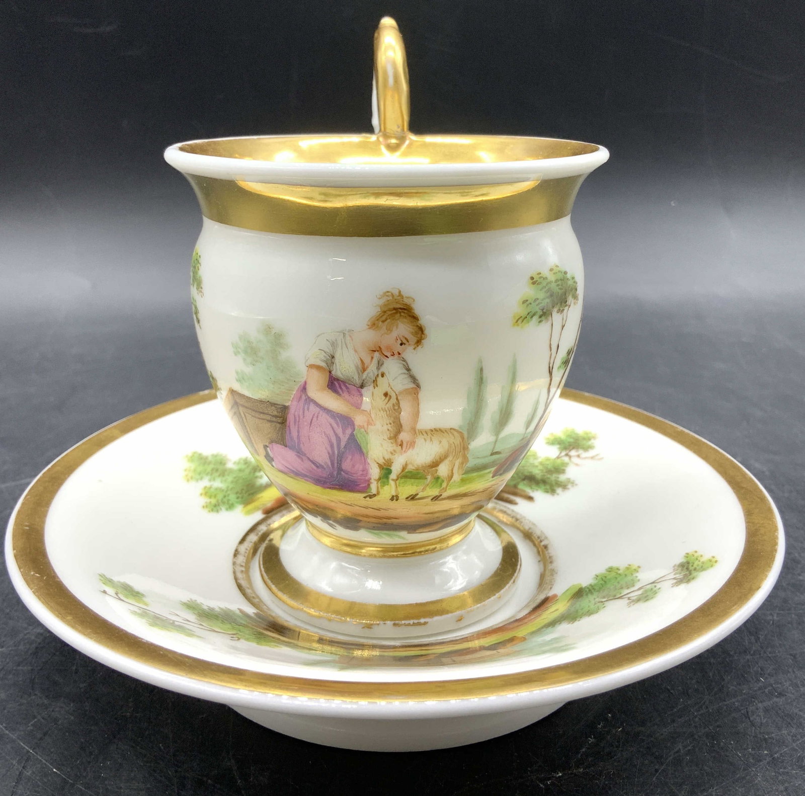 Antq VIENA Gilded Scenic Porcelain Teacup & Saucer: Antq VIENA Gilded Scenic Porcelain Teacup & Saucer Teacup & saucer featuring intricate hand-painted details, depicting a scene with a woman with a sheep & trees in the background. Edges of cups & sauc