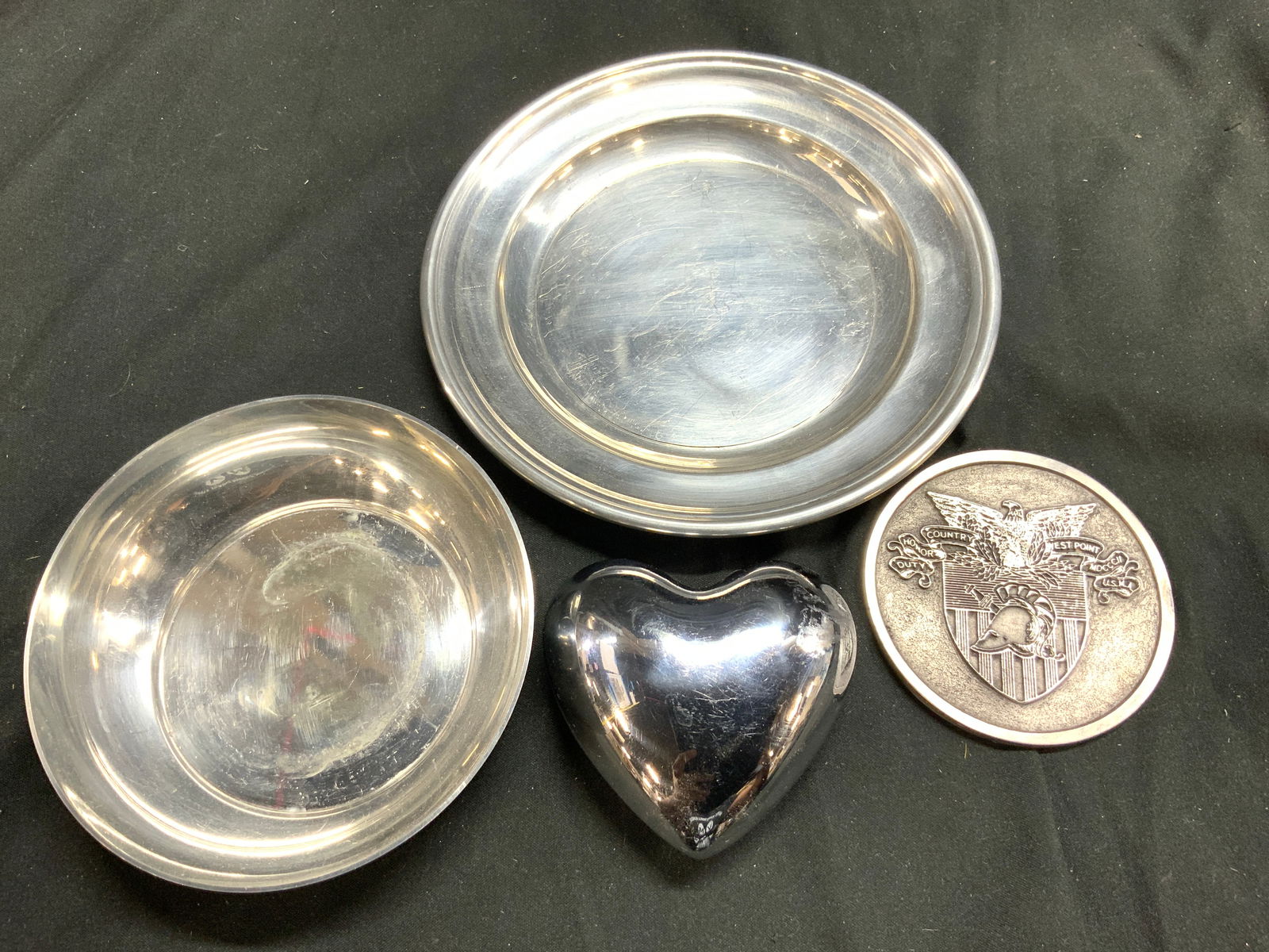Lot 4 Metal Tbltop Accs WILLIAMSBURG USA & more: Lot includes 1 tray inscribed WILLIAMSBURG, CONNECTICUT HOUSE PEWTER INC 1 WEST POINT American crest coin & 1 heart bell. Largest piece measures approximately 6 inches diameter. Metal tabletop, heart