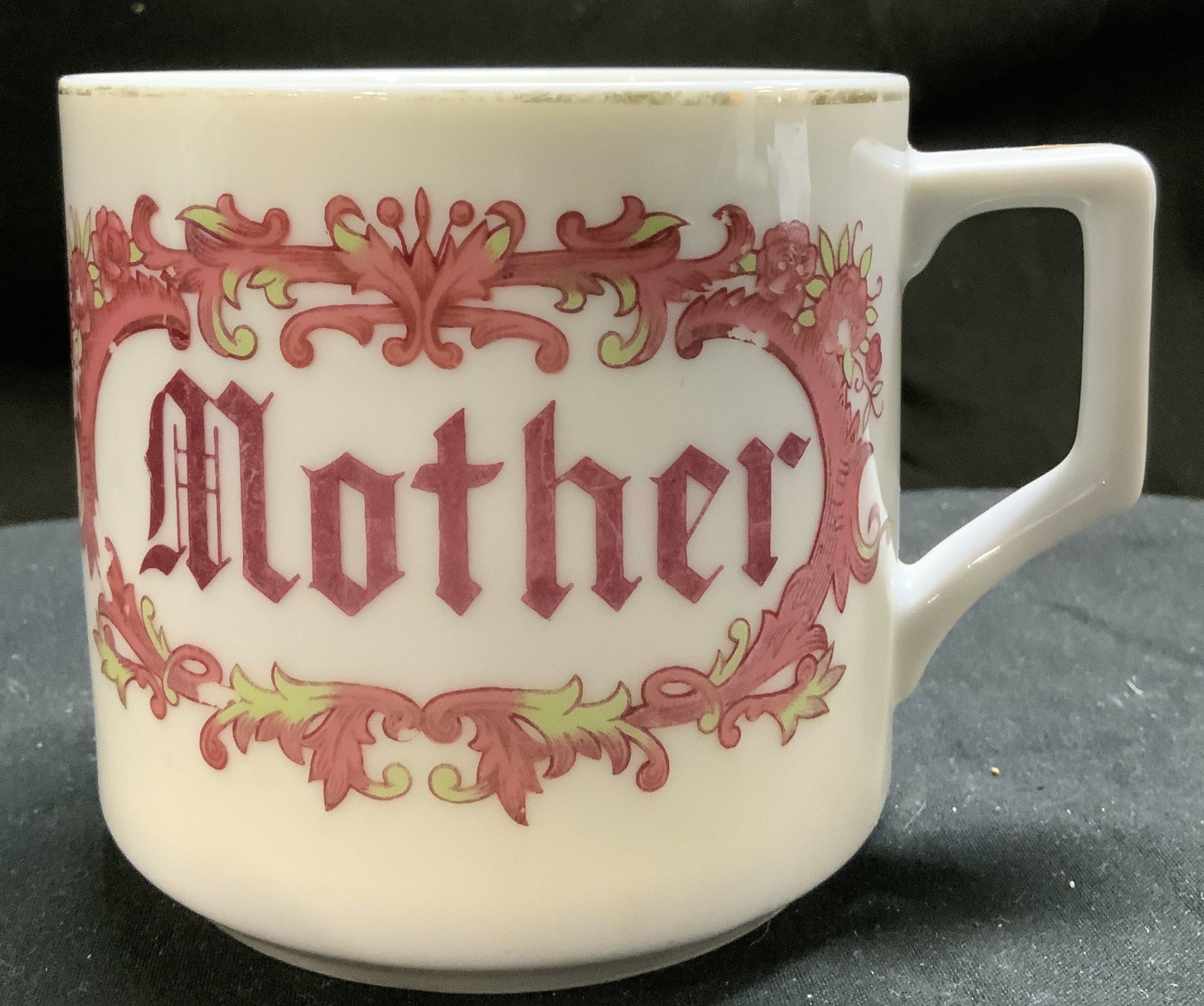 Vntg Floral Elements MOTHER Porcelain Mug: White porcelain mug featuring the word MOTHER in an ornate script. Lettering is in a pink color & surrounded by floral elements. Edges of cup & handle features faded golden accents. Surface wear consi