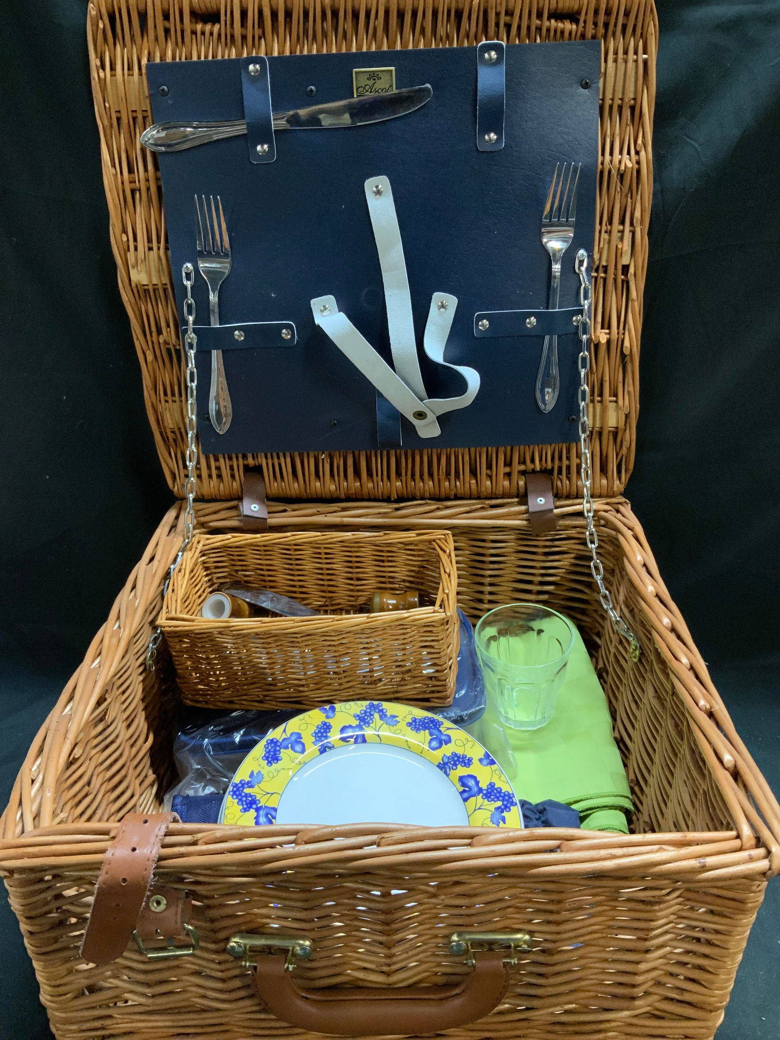 Lot 20 ASCOT Wicker Picnic Basket & Accessories: Lot includes, 2 La Primula ITALY grape motif plates, 1 small round cutting board, 1 insulated carrier from Picnic at Ascot, 1 plastic container, 2 green checkered napkins, 1 glass cup, 1 rattan storag