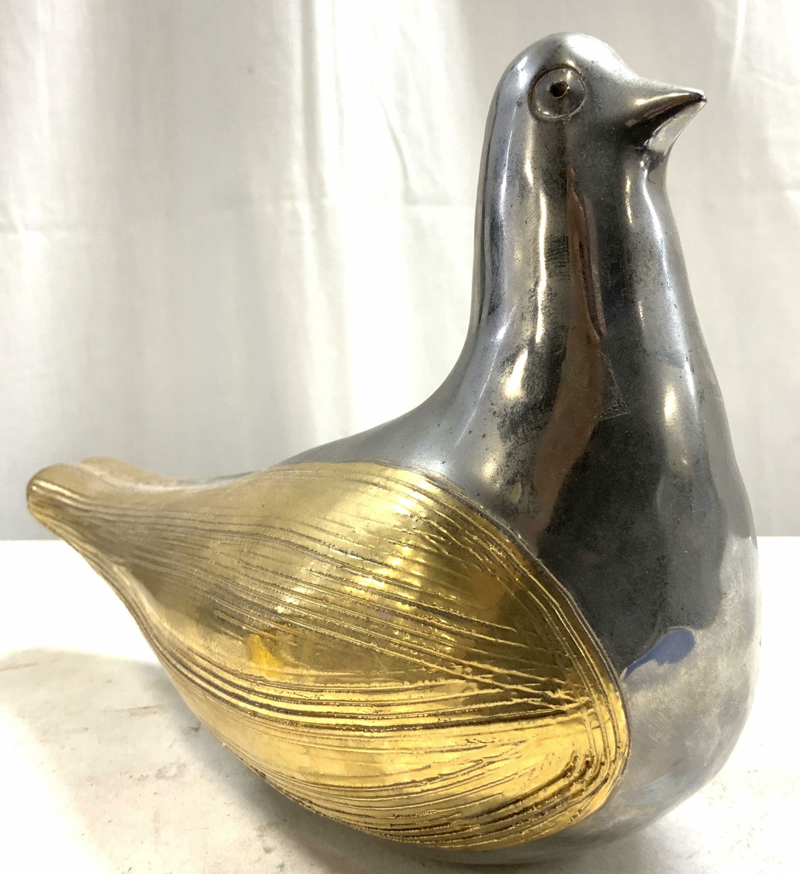 Gold & Silver toned Ceramic Pigeon Sculpture: Pigeon sculpture is ceramic. Figure is painted / glazed silver toned with gold toned wing areas with relief. Piece has relief detail on head to show eyes. Underside has tan toned unpainted area with h