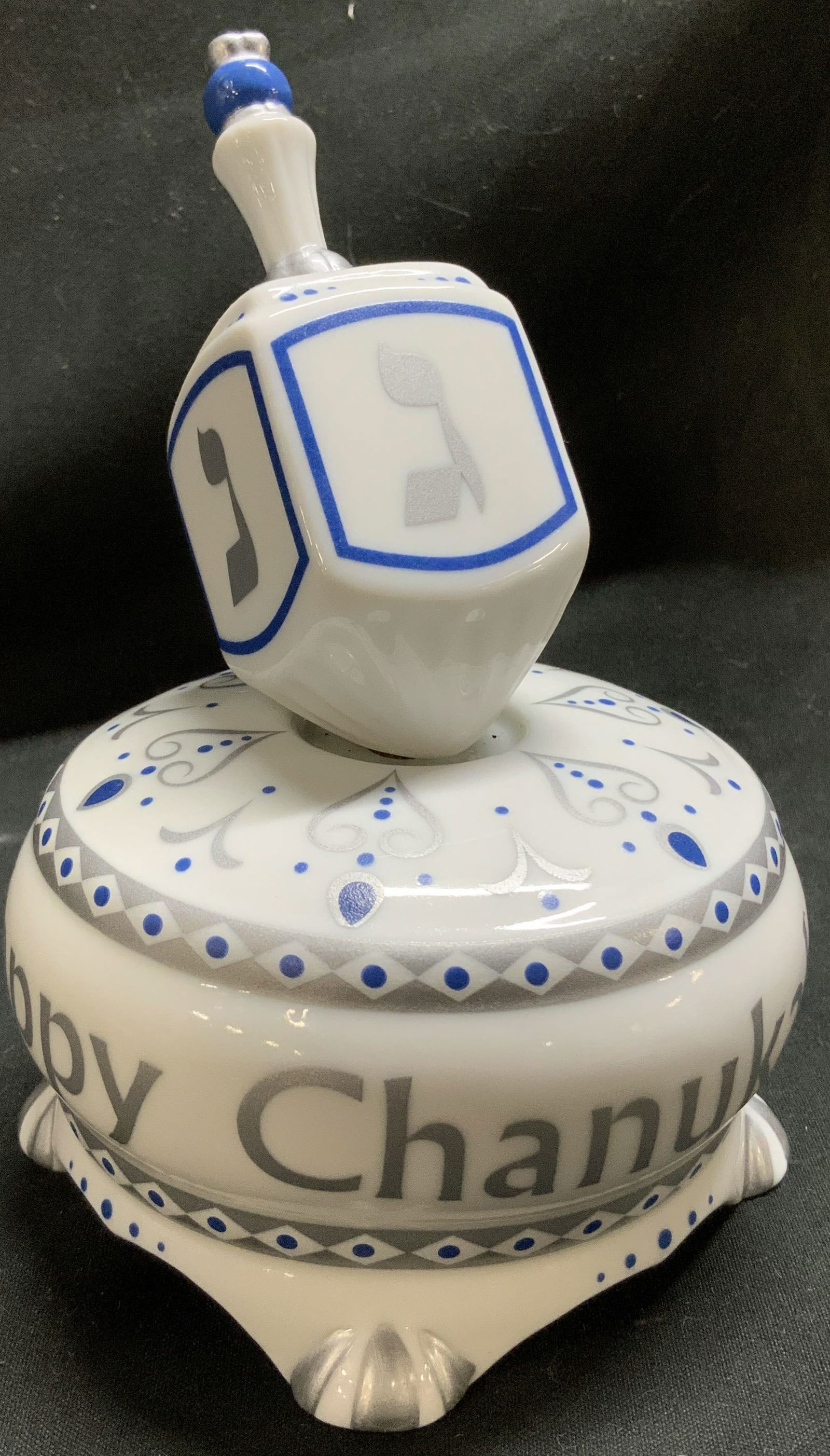Happy Chanukah Porcelain Spinning Music Box: Happy Chanukah Porcelain Spinning Music Box White and blue dreidel-shaped music box with Happy Chanukah inscribed on its base, featuring silver and blue decorative accents. Surface wear consistent to