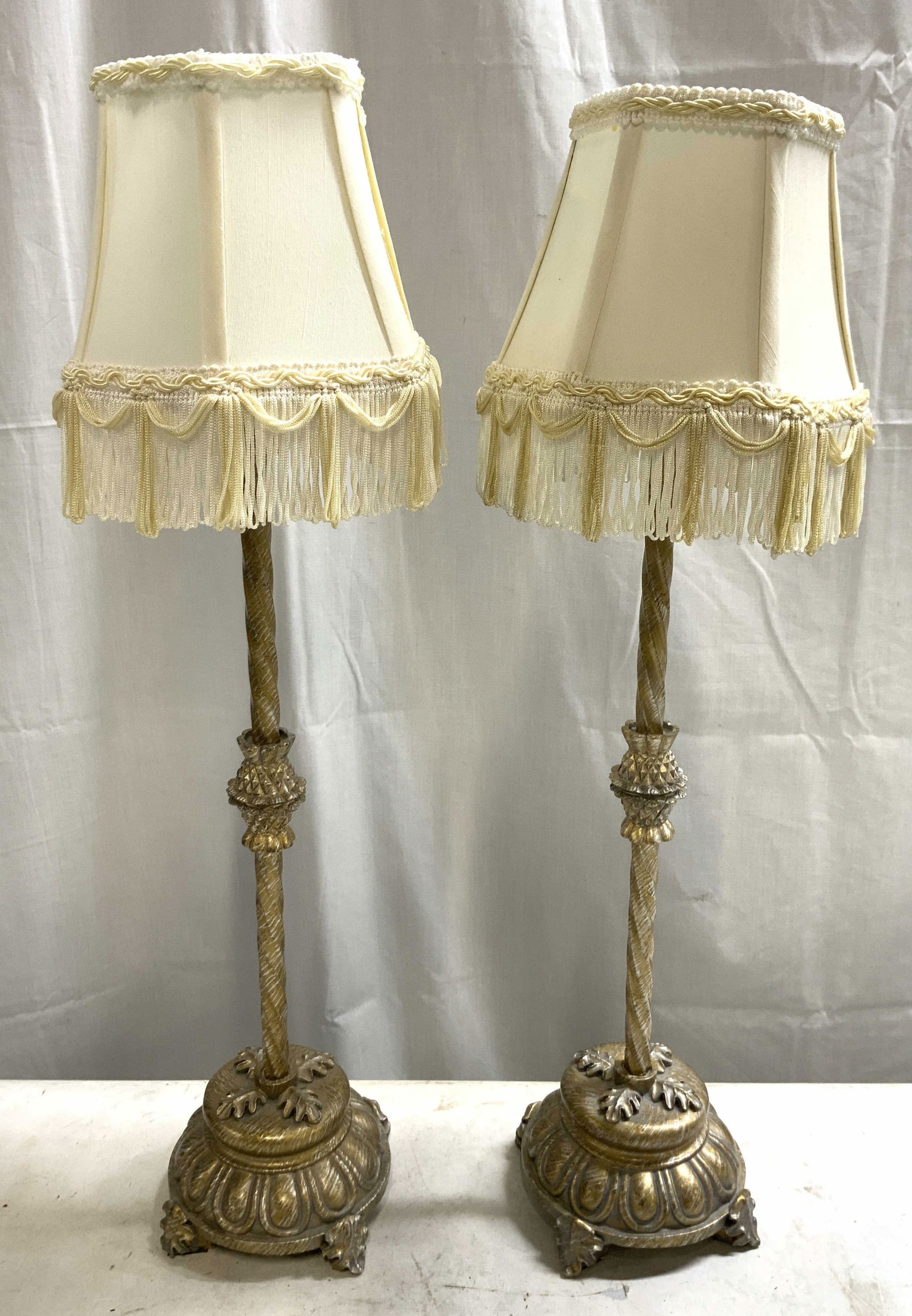 Vtg Pair Gilt Metal Tbl Lamps W Fringed Shades: Lot includes 2 lamps. Each are gilt metal. Lamps each have 4 leaf form feet. Each lamp has cream toned shades with fringes at lower edges. Each lamps measures approx 23 inches tall 5 inches wide. Lamp