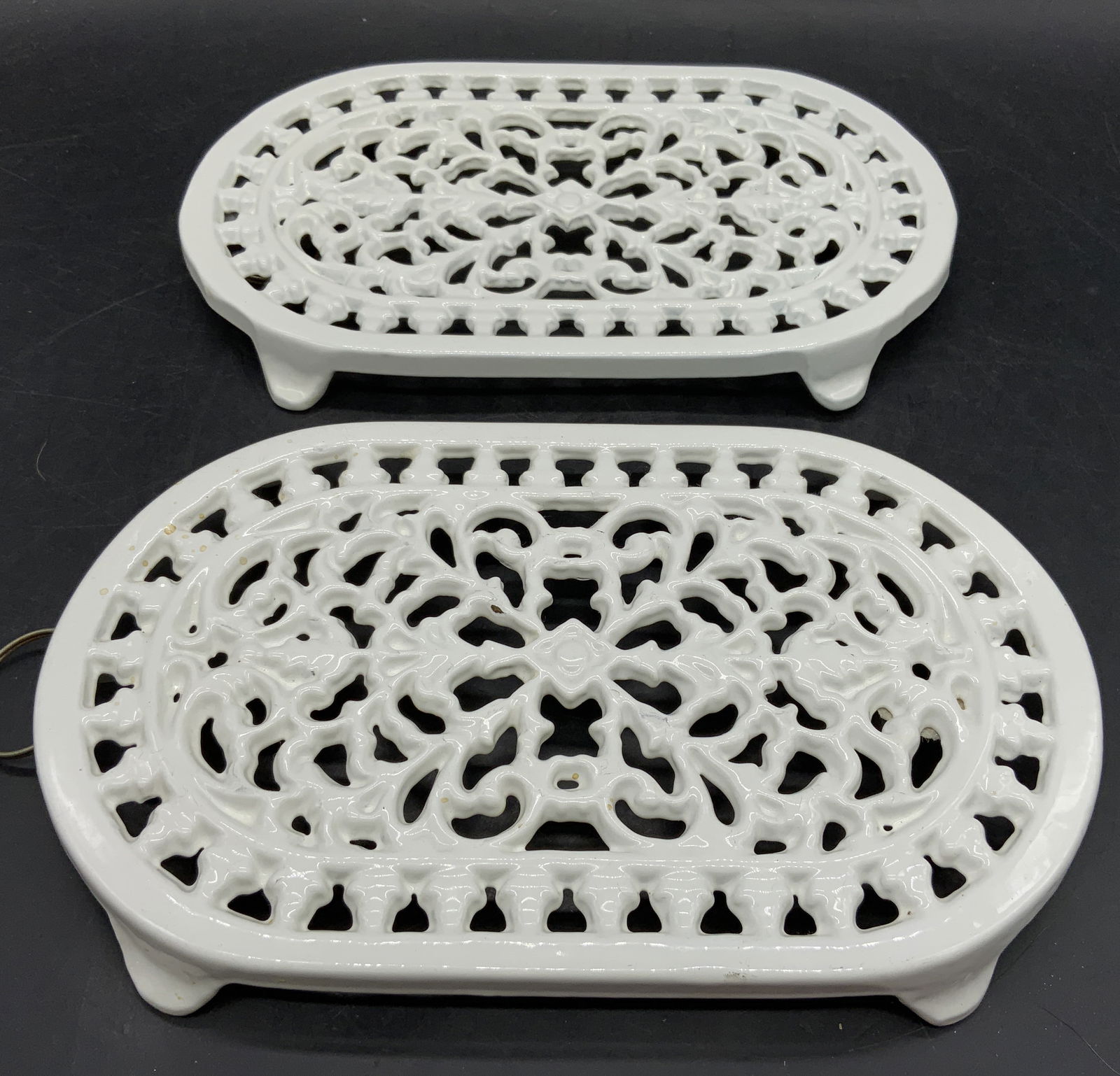 ODI Pr Enamel Iron Oval Trivets W Feet White: Undersides reads ODI. Trivets are enameled in a white tone. Each appear to be iron. Trivets are oval shaped with pierced floral motif detail. Trivets are footed. Piece measures approximately 10 inches