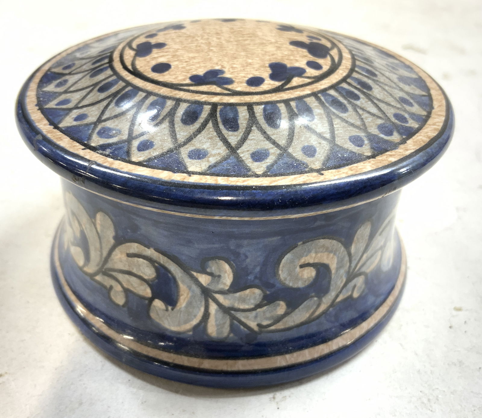 BIAGIOLI Hand Painted Trinket Bx GUBBIO ITALY: Underside is signed BIAGIOLI GUBBIO ITALY. Trinket box is hand painted. Piece appears to be ceramic. Box is circular, tan with blue toned scroll and leaf detail. Trinket box measures approximately 3.7