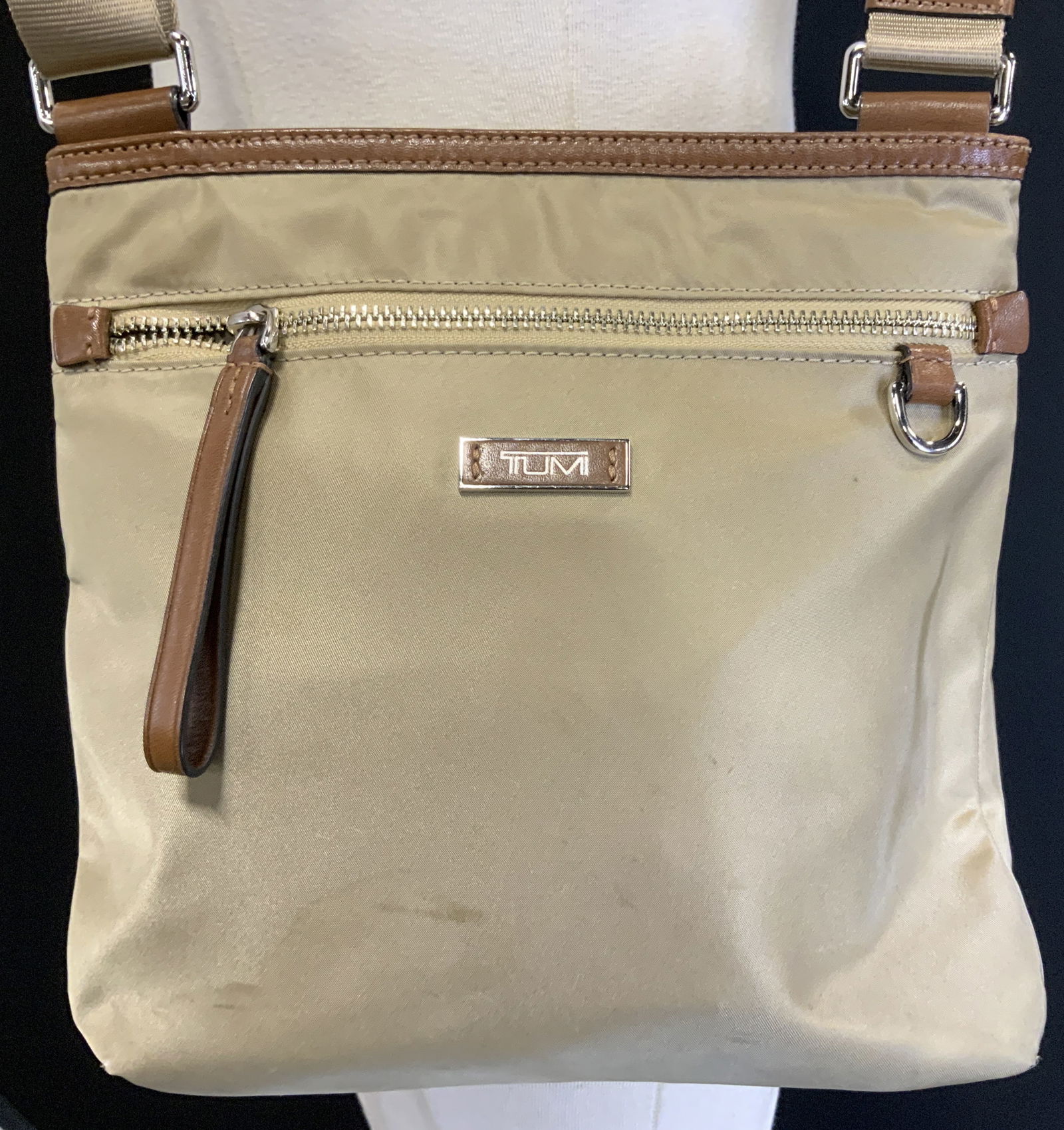Vntg Tumi Voyageur Canton Beige Crossbody Bag: The bag features brown leather trim and accents, including a zipper pull and strap attachments. Center of bag features a metal plaque that reads, TUMI. Signed & marked. Reads, TUMI. Surface wear consi