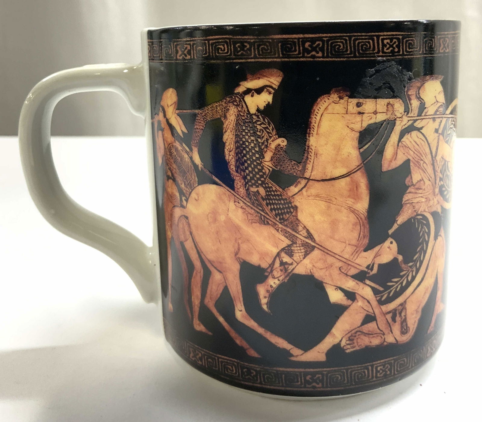 THE MET Trojan Style Porcelain Mug: Underside reads THE MET. Mug is porcelain. Piece is black with detail depicting Trojan soldiers and horses in battle. Mug has white toned handle and interior. Measures approximately 3.75 inches tall 4