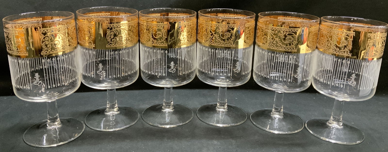 Set 6 Vntg Culver Starlyte Footed Glass Goblets: Footed clear glass goblet with a stem. The upper part of the glass features a wide, ornate gold band with a patterned design. Below the gold band, the glass is decorated with a pattern of thin white v