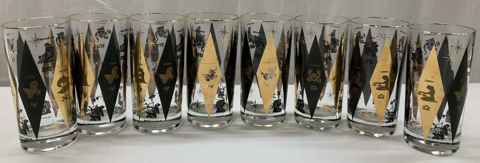 Set8 Harlequin Zodiac Highball Glass Tumblers: Highball glass tumblers feature a black and gold harlequin diamond pattern that includes the astrological signs. Signs include, Aries, Pisces, Taurus, Virgo, Sagittarius, Libra, Cancer, Capricorn. Gla