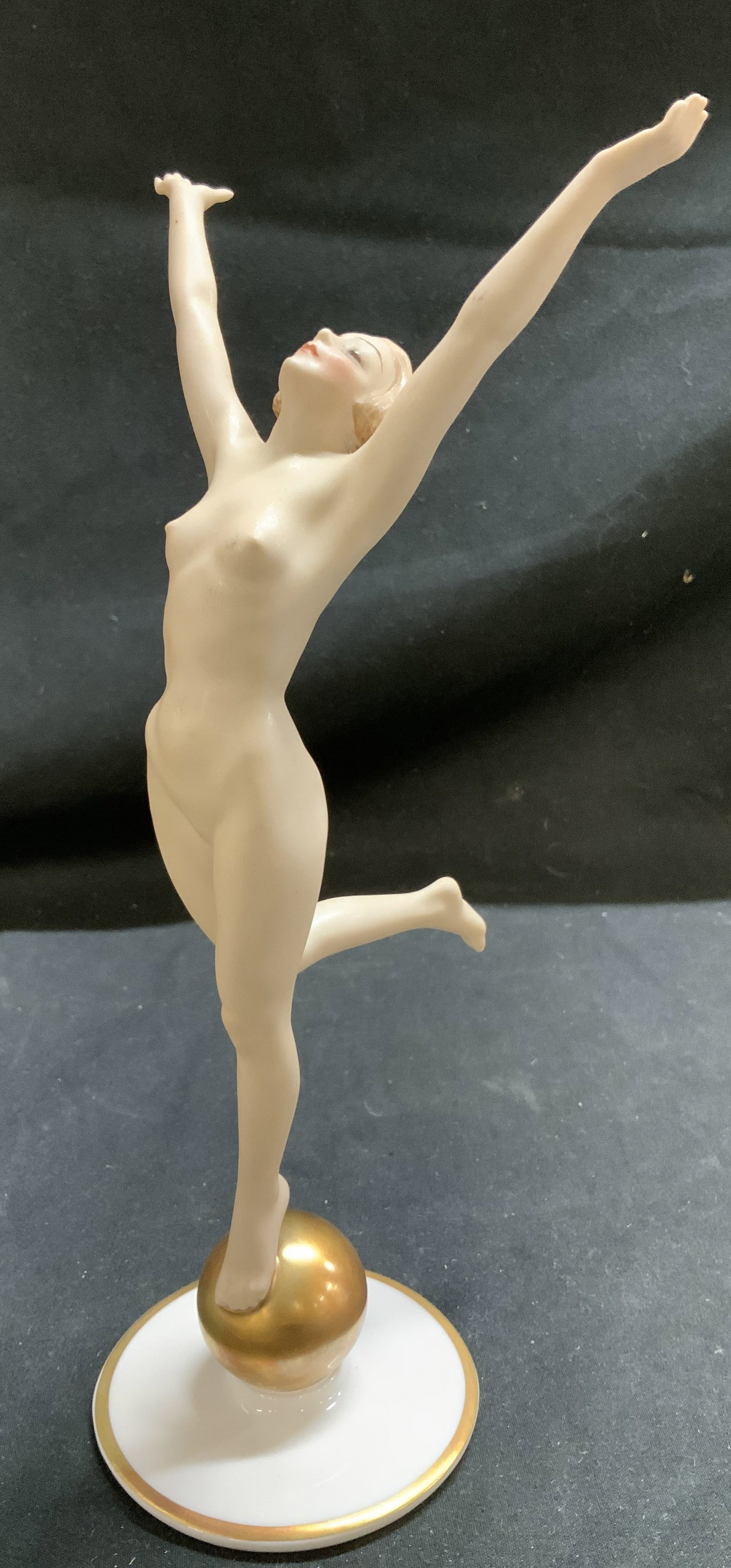Vntg Hutschenreuther Sonnenkind Porc Fig. GERMANY: Porcelain figurine depicting nude female with outstretched arms and one leg raised, balancing on a golden ball. Designed by Karl Tutter around 1930. Figurine features gold accents on base. Surface wea
