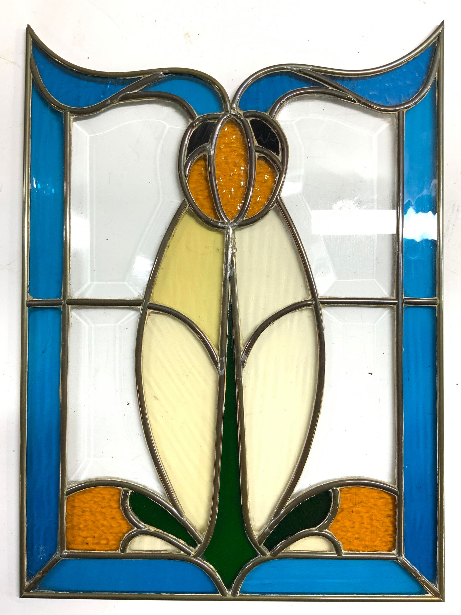 Floral Stained Glass Art Glass Wall Hanging: Wall hanging is stained glass piece has floral detail. Clear glass areas are beveled. Piece has blue toned edges. Center of piece has green, orange, yellow and black tones. Stained glass wall hanging