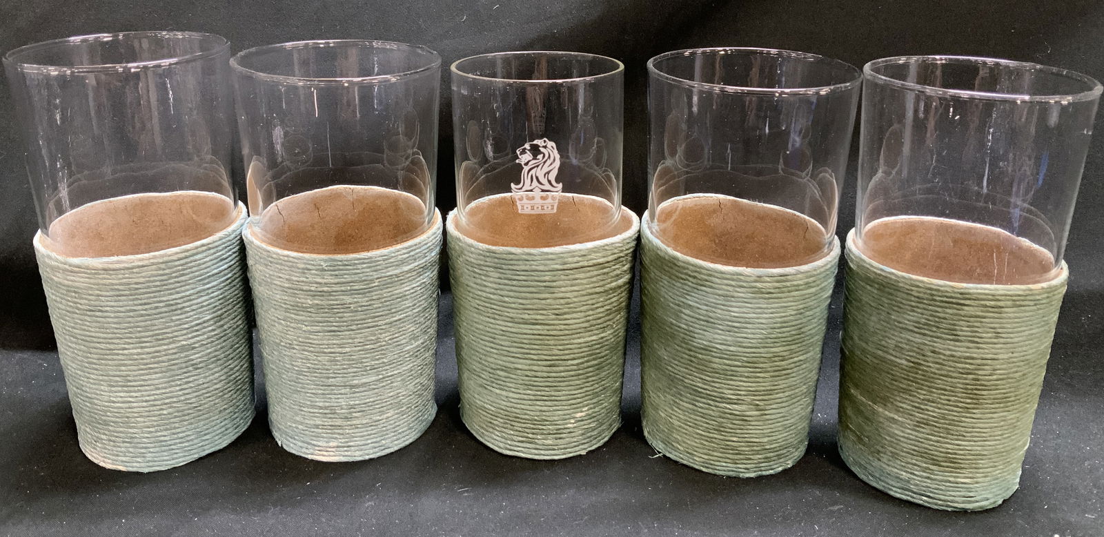 Lot 5 Clear Glass Tumblers W Twine Wrapped Base: Lot 5 Clear Glass Tumblers W Twine Wrapped Base Lot includes 5 clear class tumblers featuring a green twine base. 1 tumbler features a crowned lion emblem. Surface wear consistent to age, use & exposu