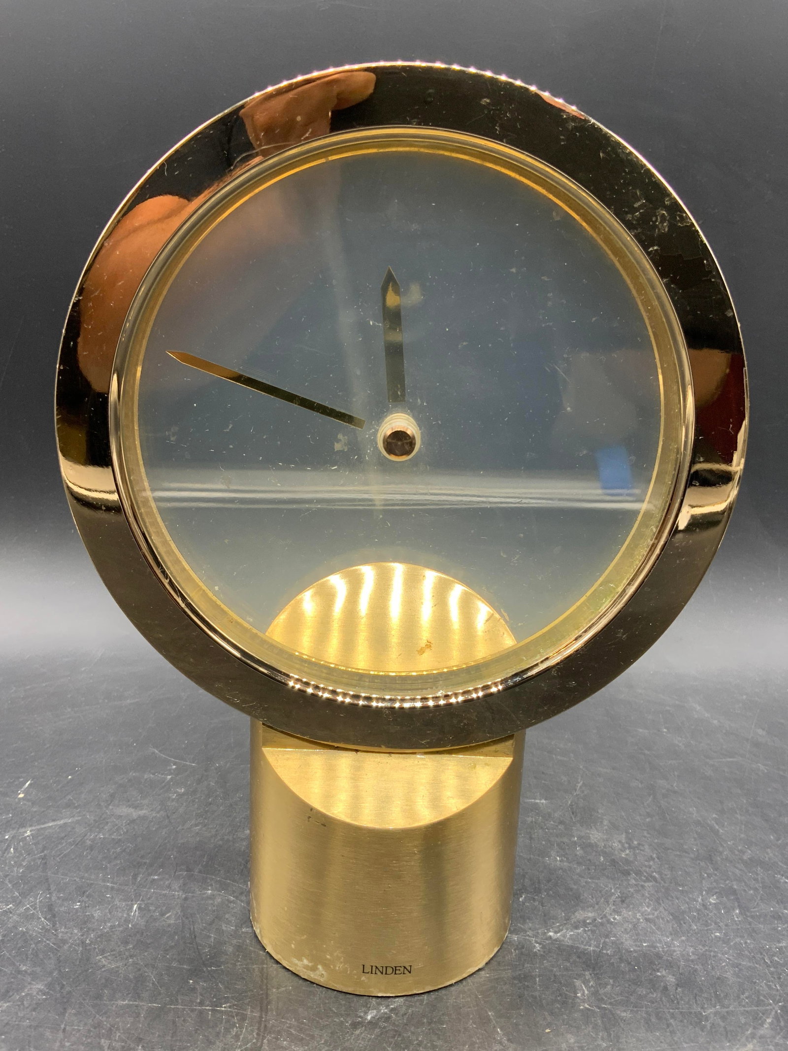 LINDEN Minimalist MYSTERY Tabletop Clk JAPAN: Clock base is trademarked LINDEN. Clock is gold toned brushed metal, appears to be brass with minimalist glass face and gold toned hands. Underside is black toned & Reads JAPAN. Clock measures approxi